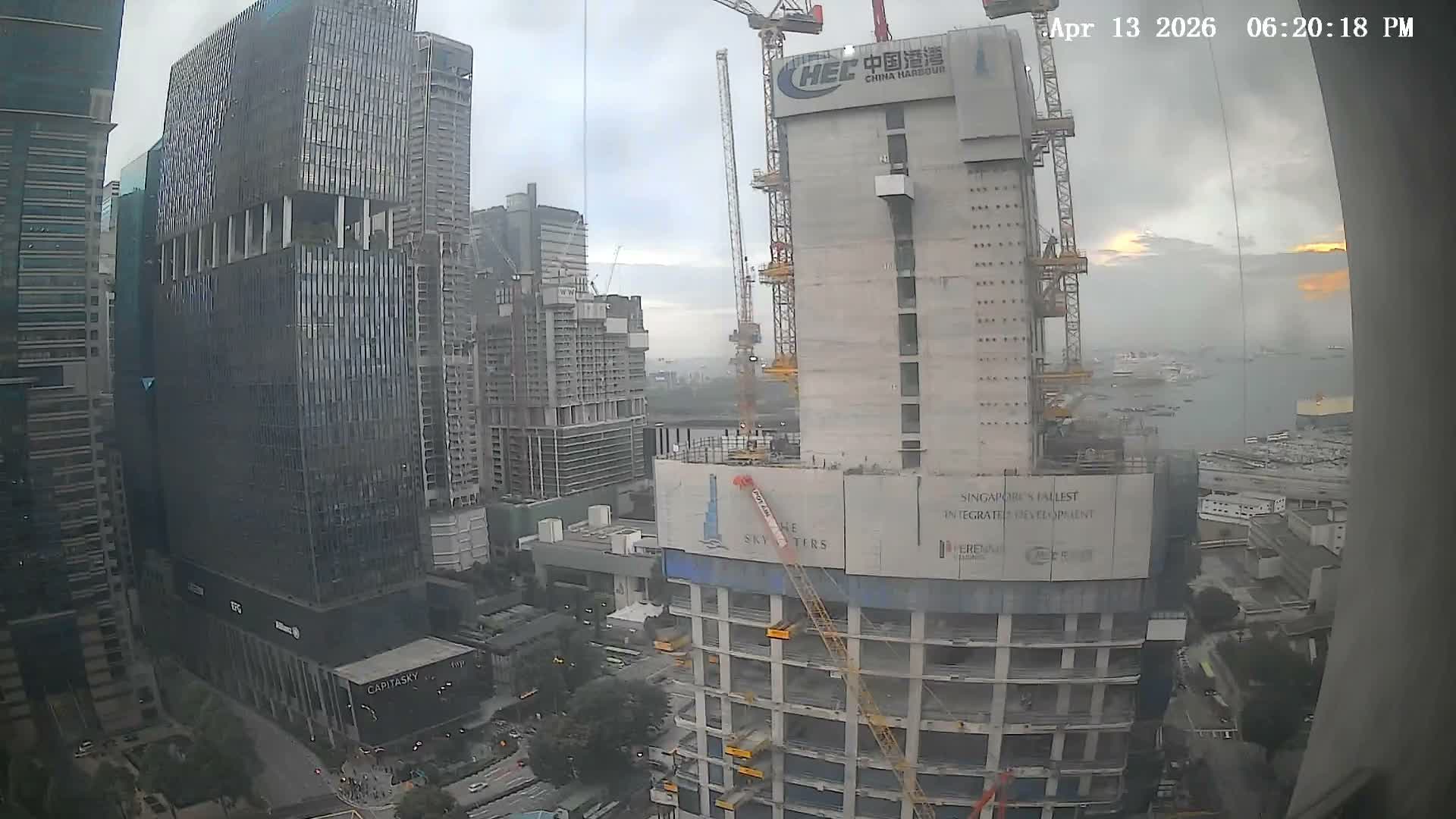 Numerous skyscrapers and active construction sites with multiple cranes dominate an urban landscape that overlooks a busy harbor filled with ships under a partly cloudy sky.