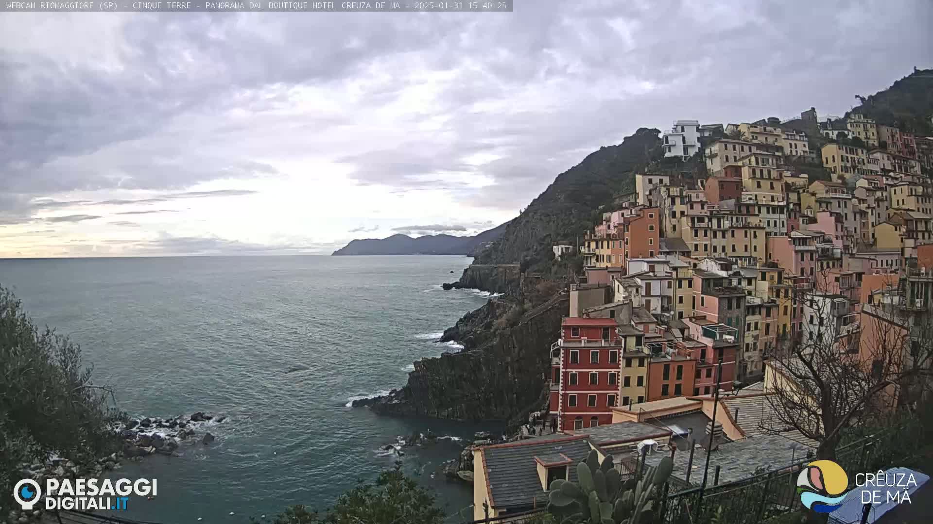 A coastal town with colorful buildings clinging to a steep cliff overlooking a calm sea under a cloudy sky.