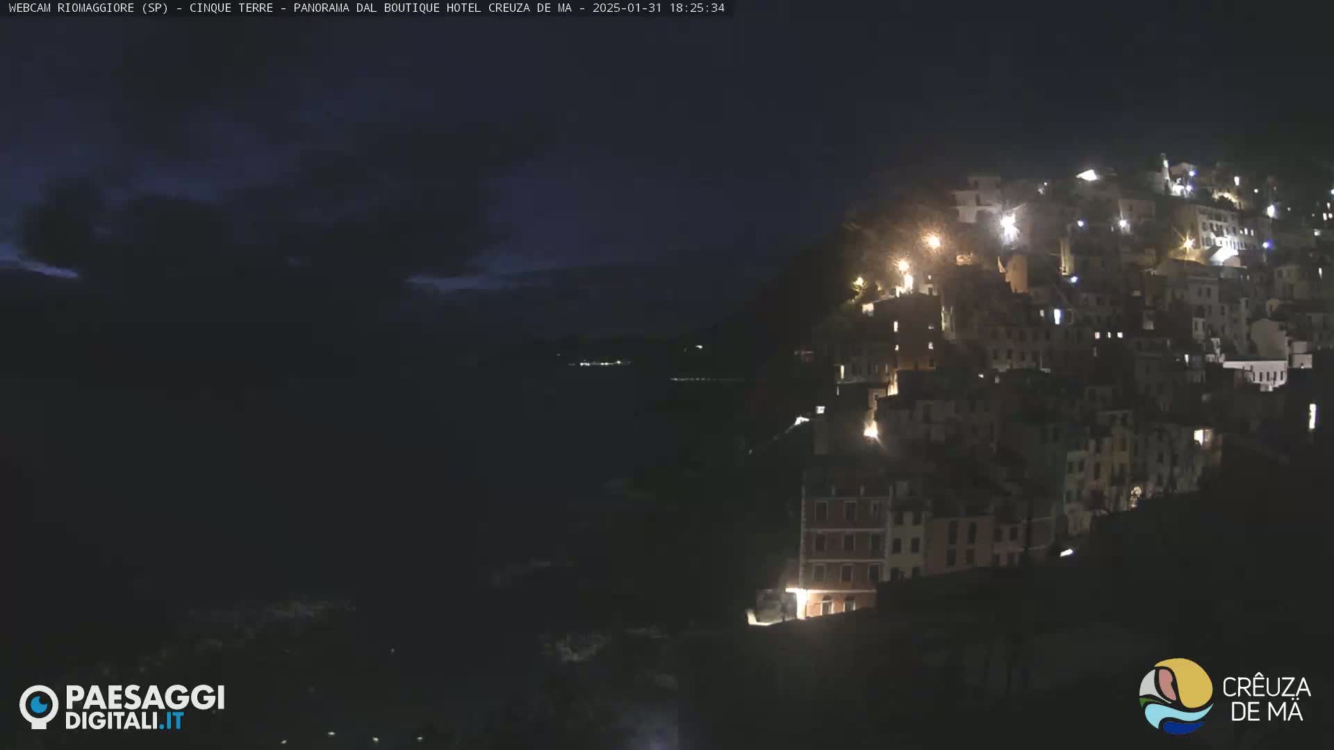 A nighttime view of a hillside village with illuminated buildings overlooking a dark body of water under a partly cloudy sky.