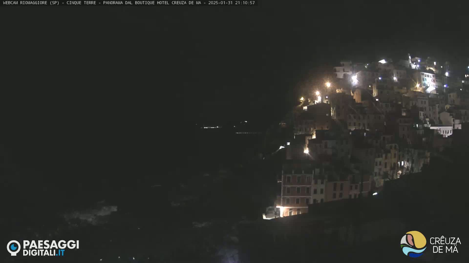 A nighttime view of a hillside town illuminated by scattered lights, overlooking a dark body of water.