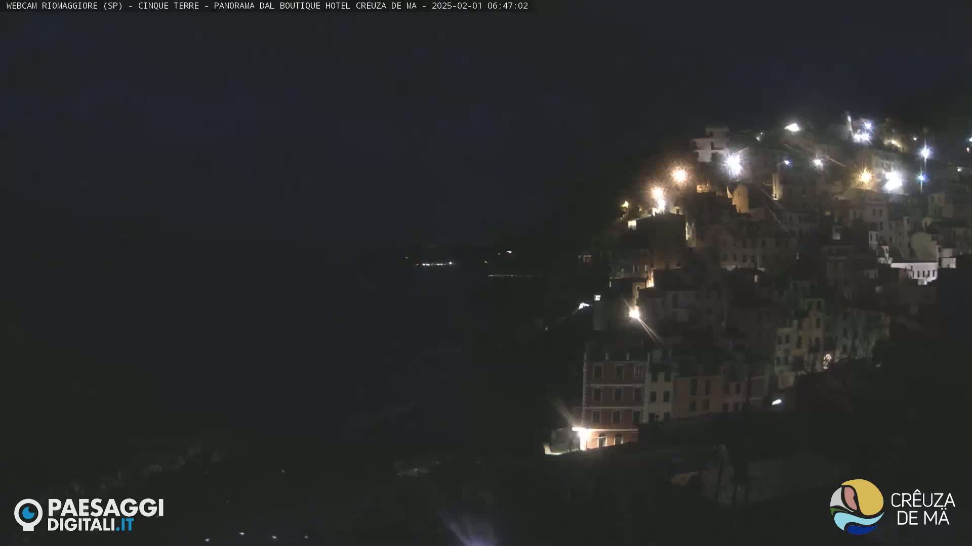 A nighttime, long shot of a hillside town illuminated by scattered lights, overlooking a dark body of water.
