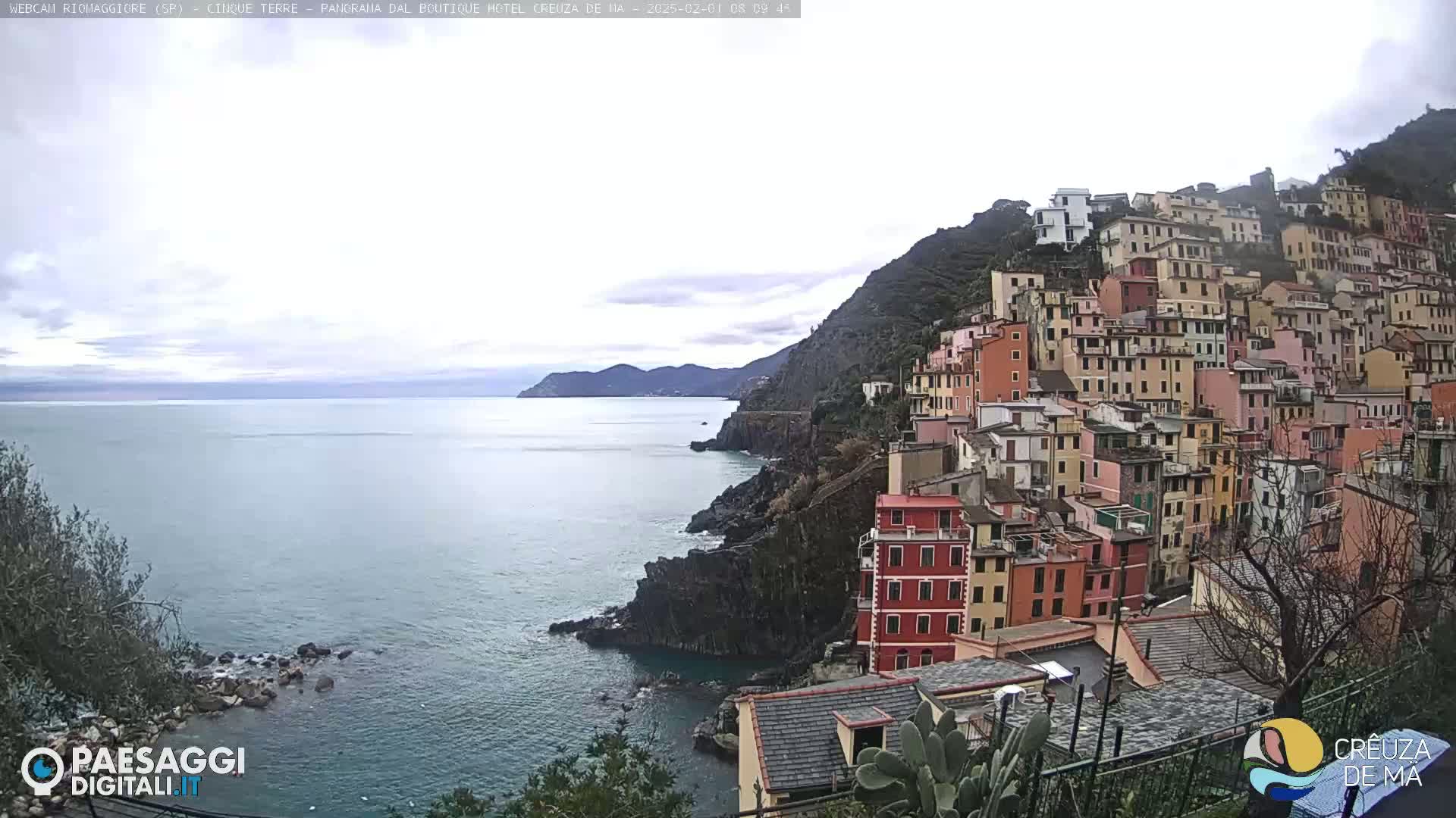 A coastal town of colorful buildings on a hillside overlooks a calm sea under an overcast sky.