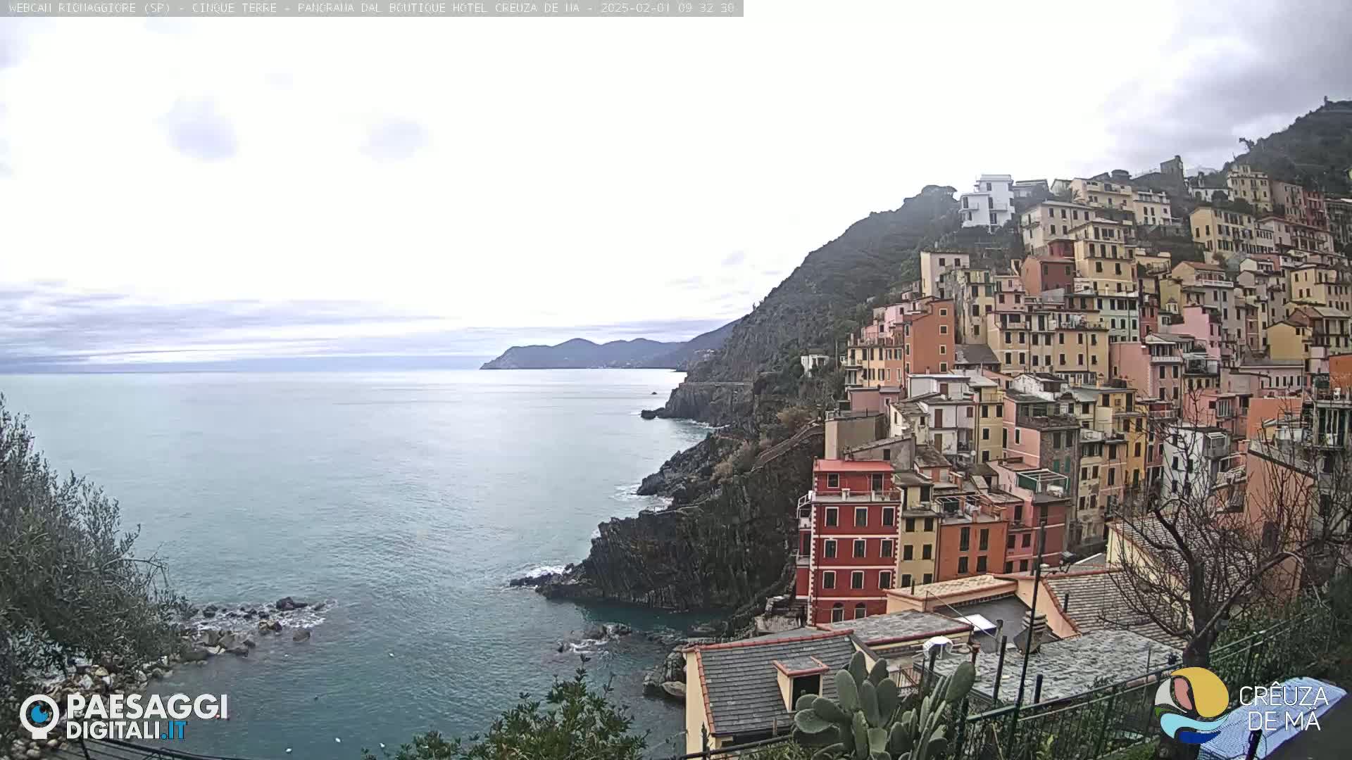 A coastal town with colorful buildings stacked on a hillside overlooks a calm sea under an overcast sky.