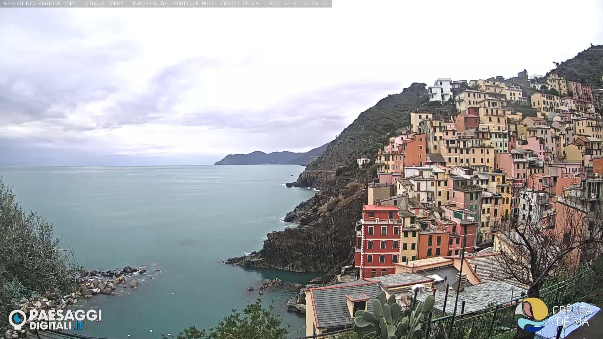 A colorful village clings to a steep, rocky hillside overlooking a calm, blue-green sea under an overcast sky.