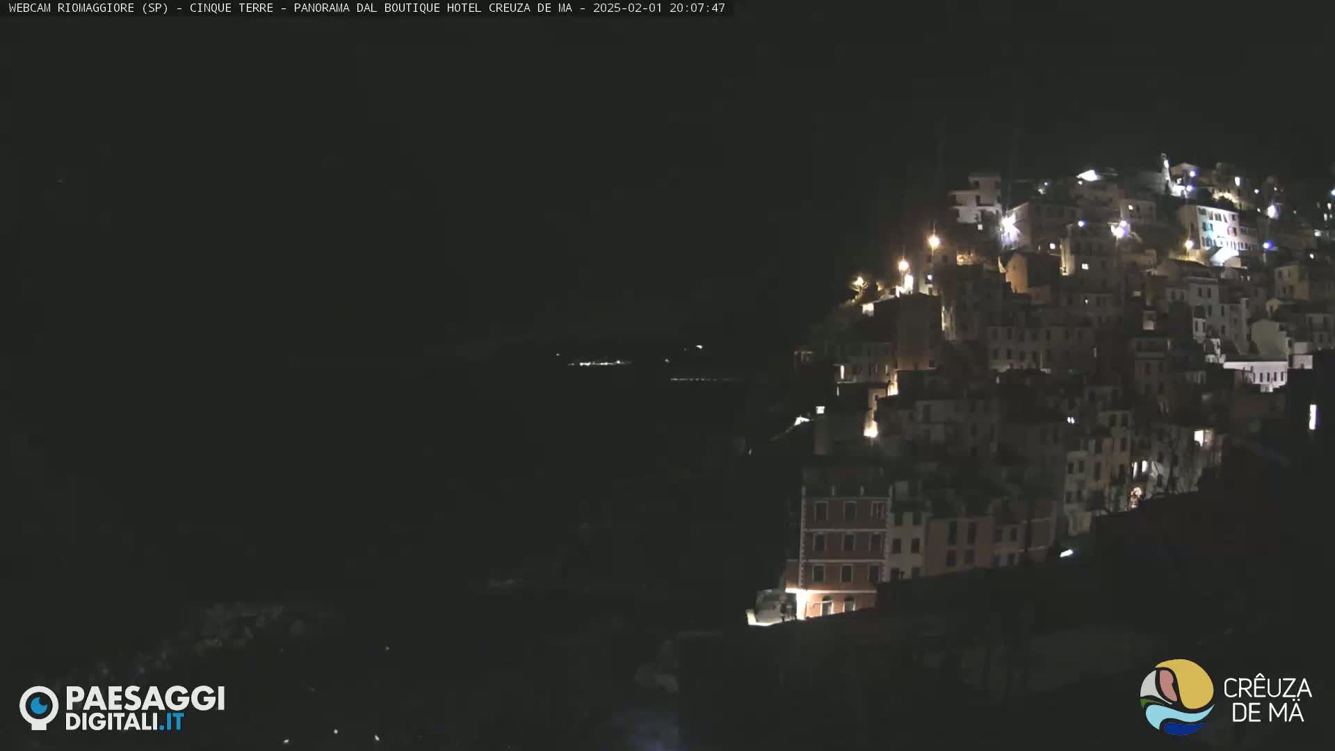 A nighttime view of a hillside town illuminated by lights, overlooking a dark body of water.