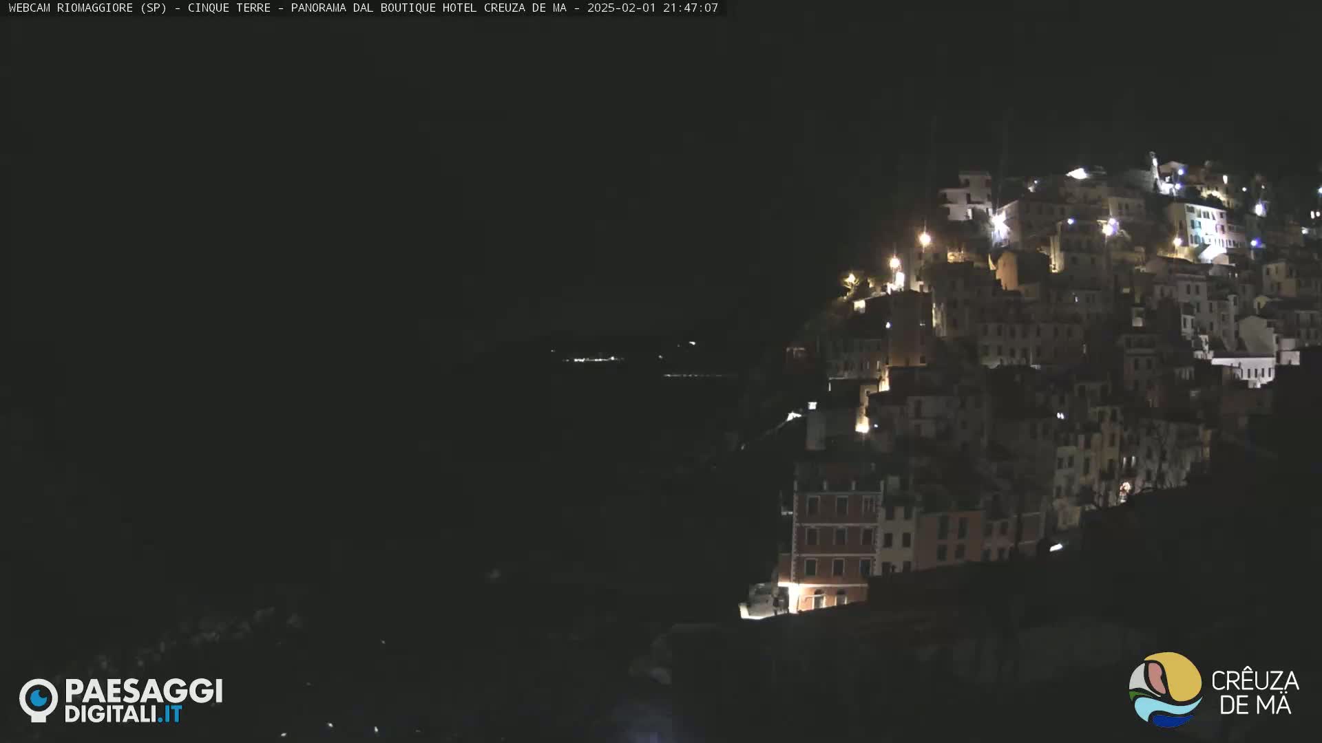 A nighttime view of a hillside town illuminated by scattered lights, overlooking a dark body of water.