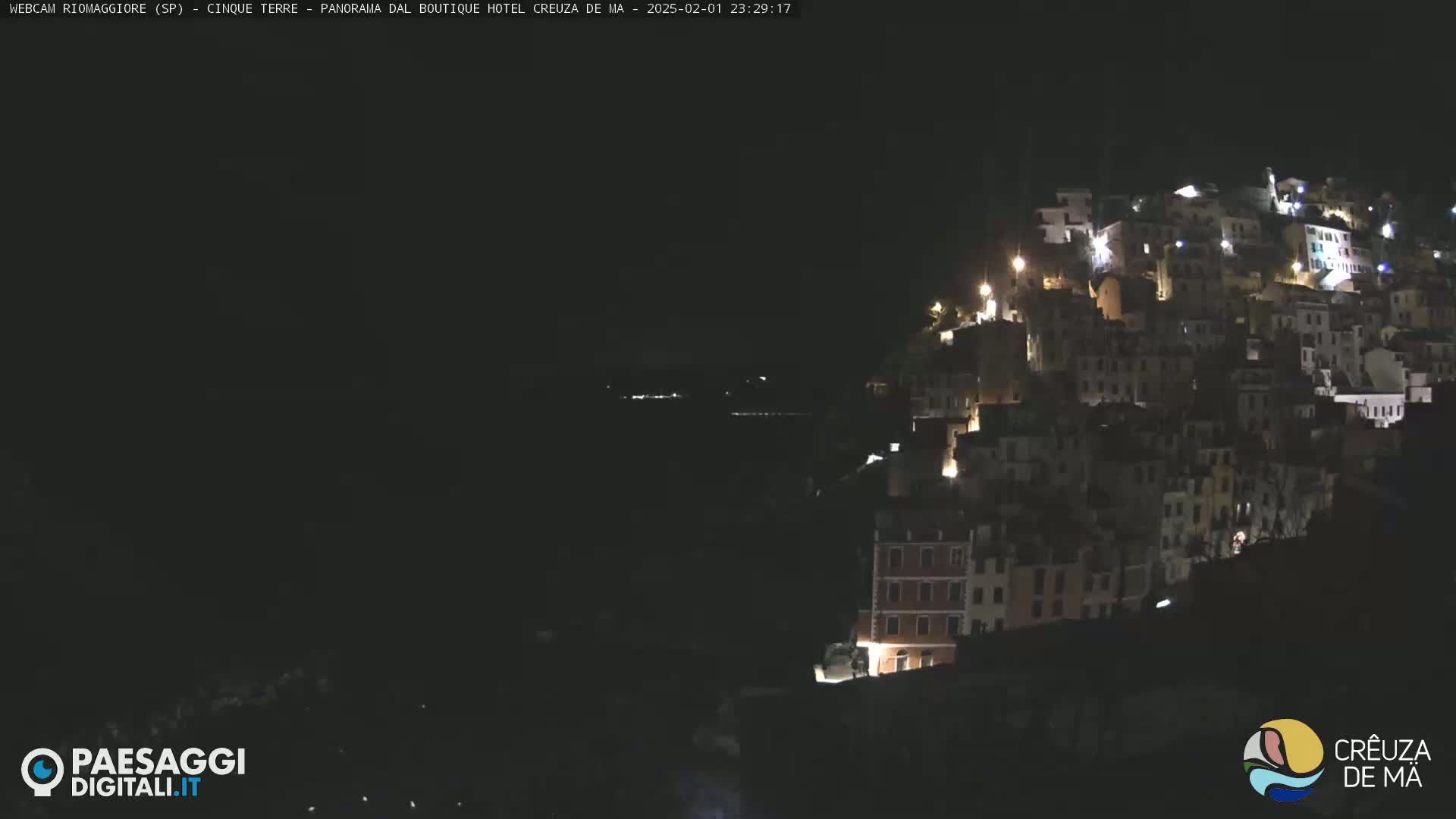 A nighttime view of a hilltop town illuminated by scattered lights, overlooking a dark body of water.