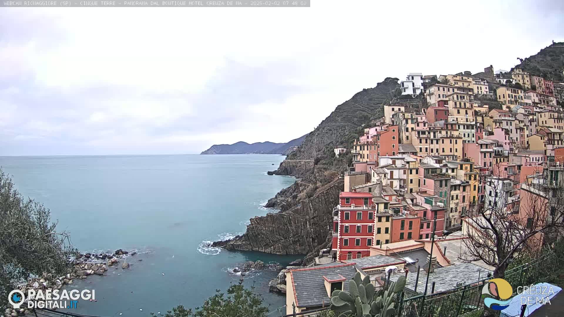 A coastal town of colorful buildings clings to a steep hillside overlooking a calm, blue-green sea under an overcast sky.