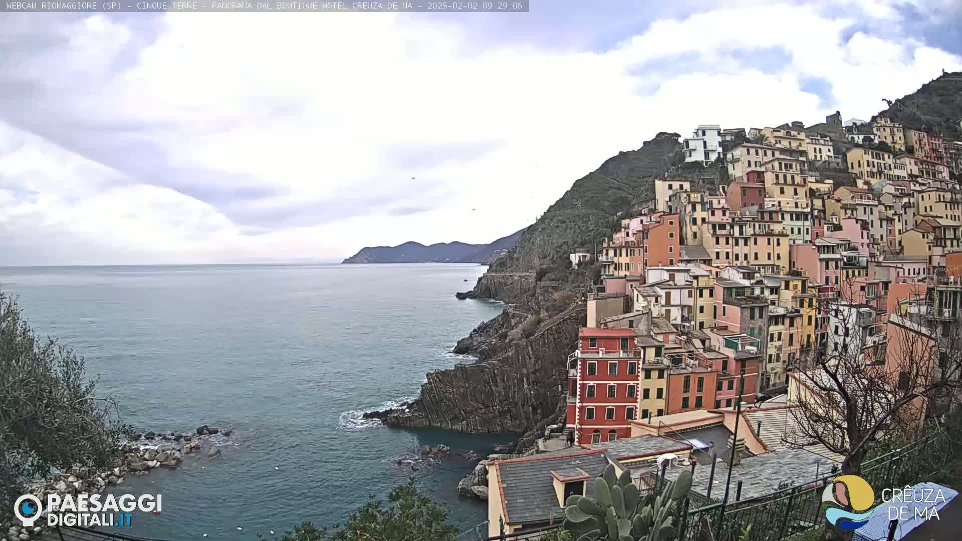 A coastal village of colorful buildings climbs a steep hillside overlooking a calm sea under a partly cloudy sky.