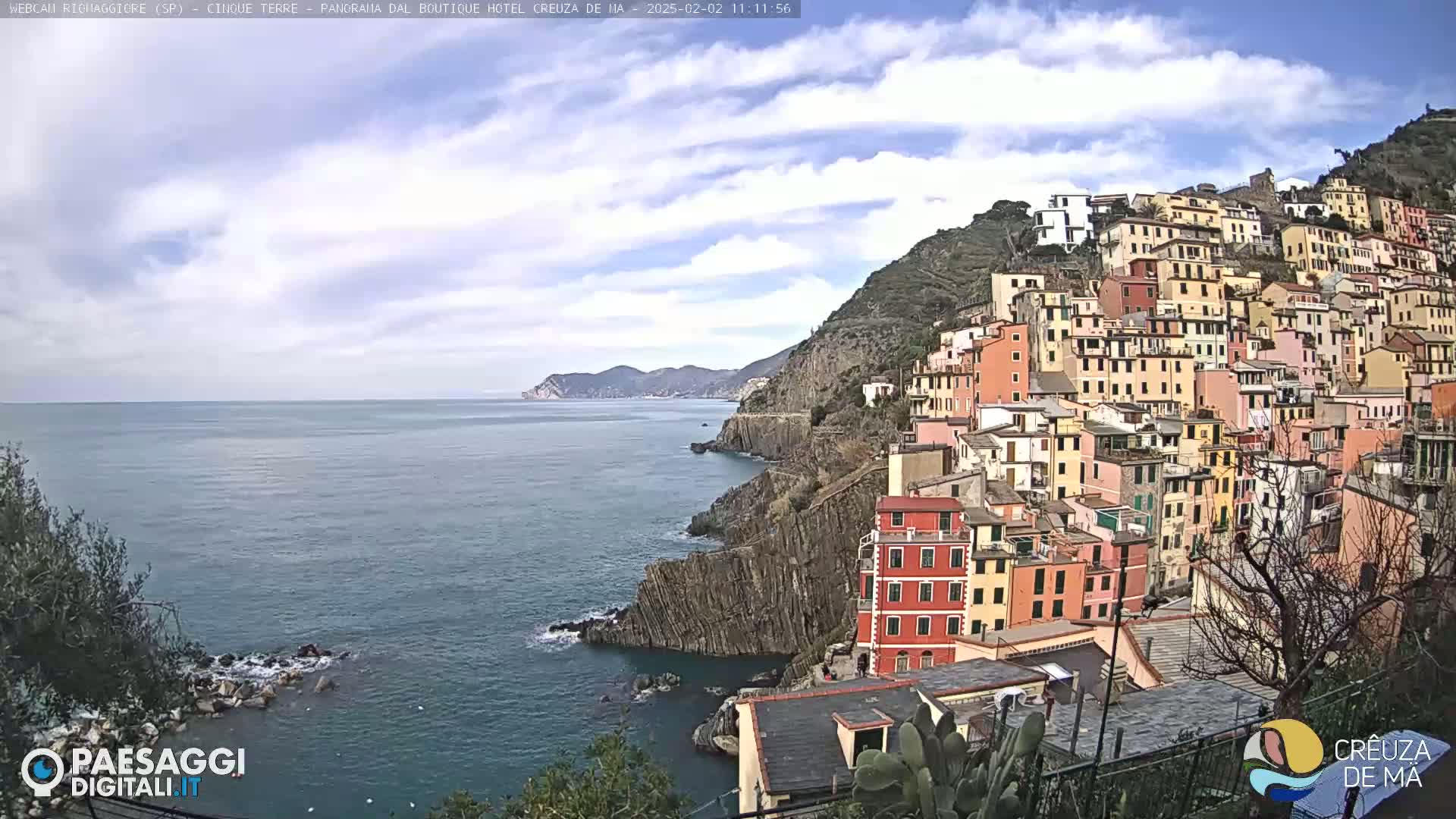 A colorful hillside village clings to a rocky coastline overlooking a calm sea under a partly cloudy sky.