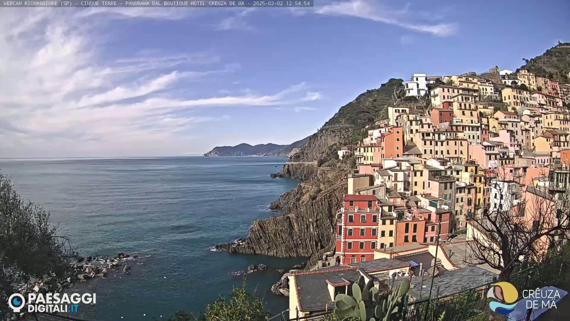A colorful hillside village clings to a rocky coast overlooking a calm sea under a mostly clear blue sky.