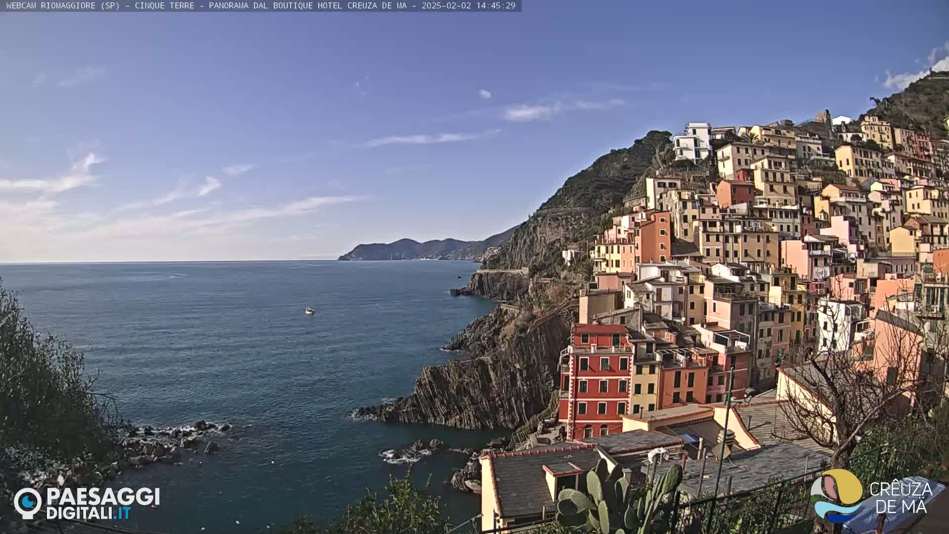A colorful village clings to a steep, rocky hillside overlooking a calm blue sea under a mostly clear sky.