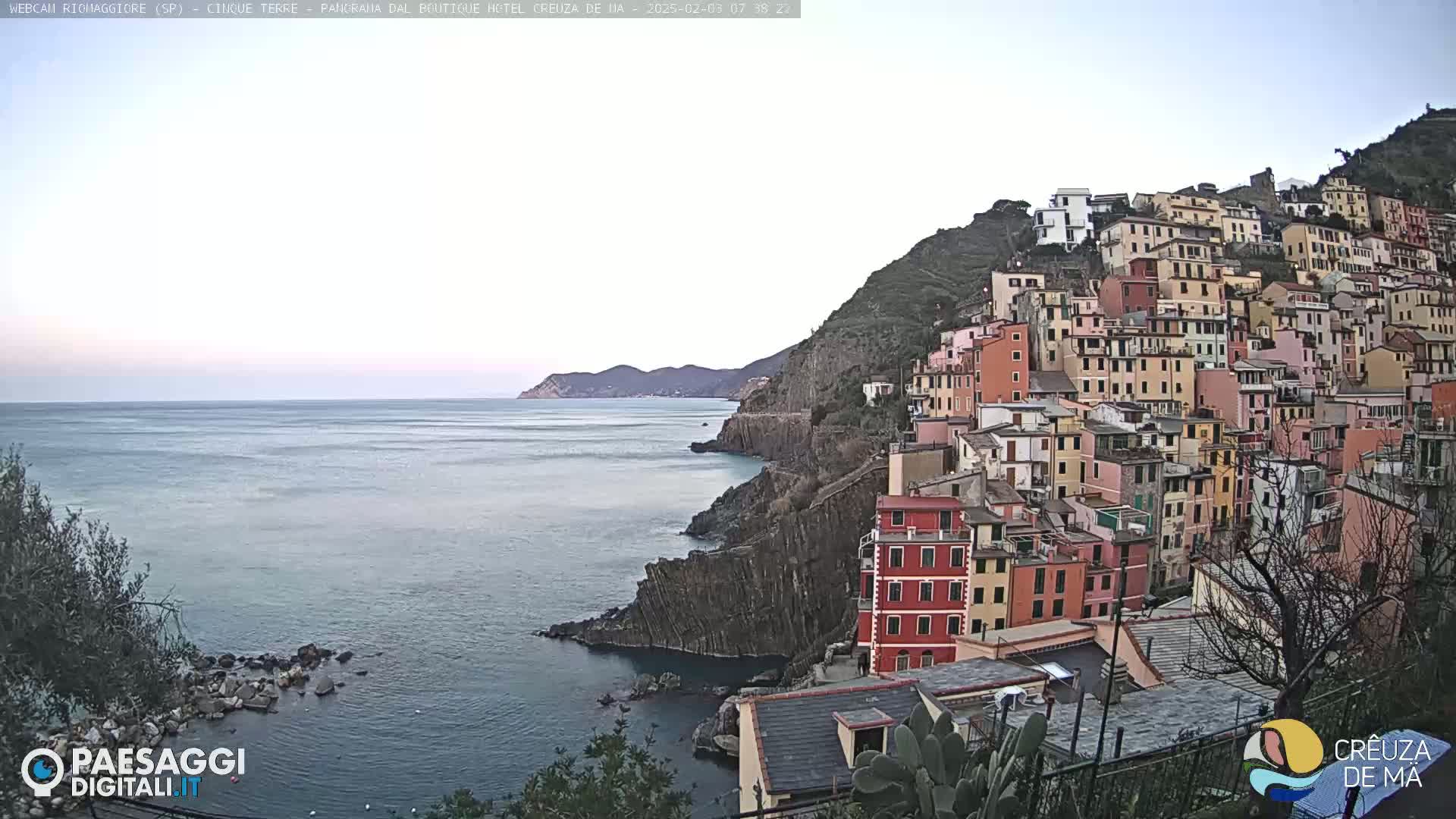 A coastal town with colorful buildings clinging to a steep hillside overlooks a calm sea under a clear, pale sky.