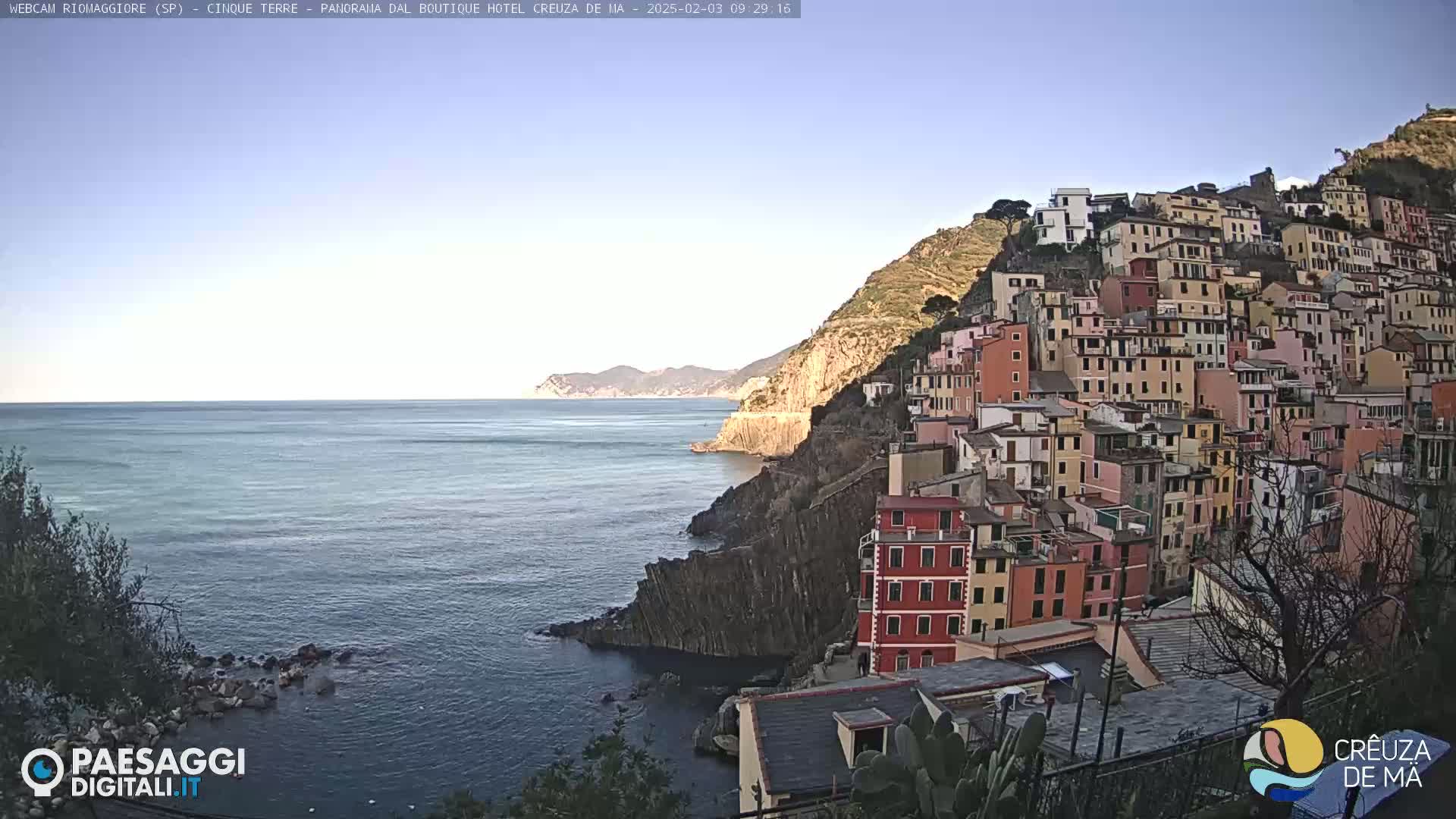 A colorful hillside village clings to a rocky coastline overlooking a calm, blue sea under a clear, bright sky.