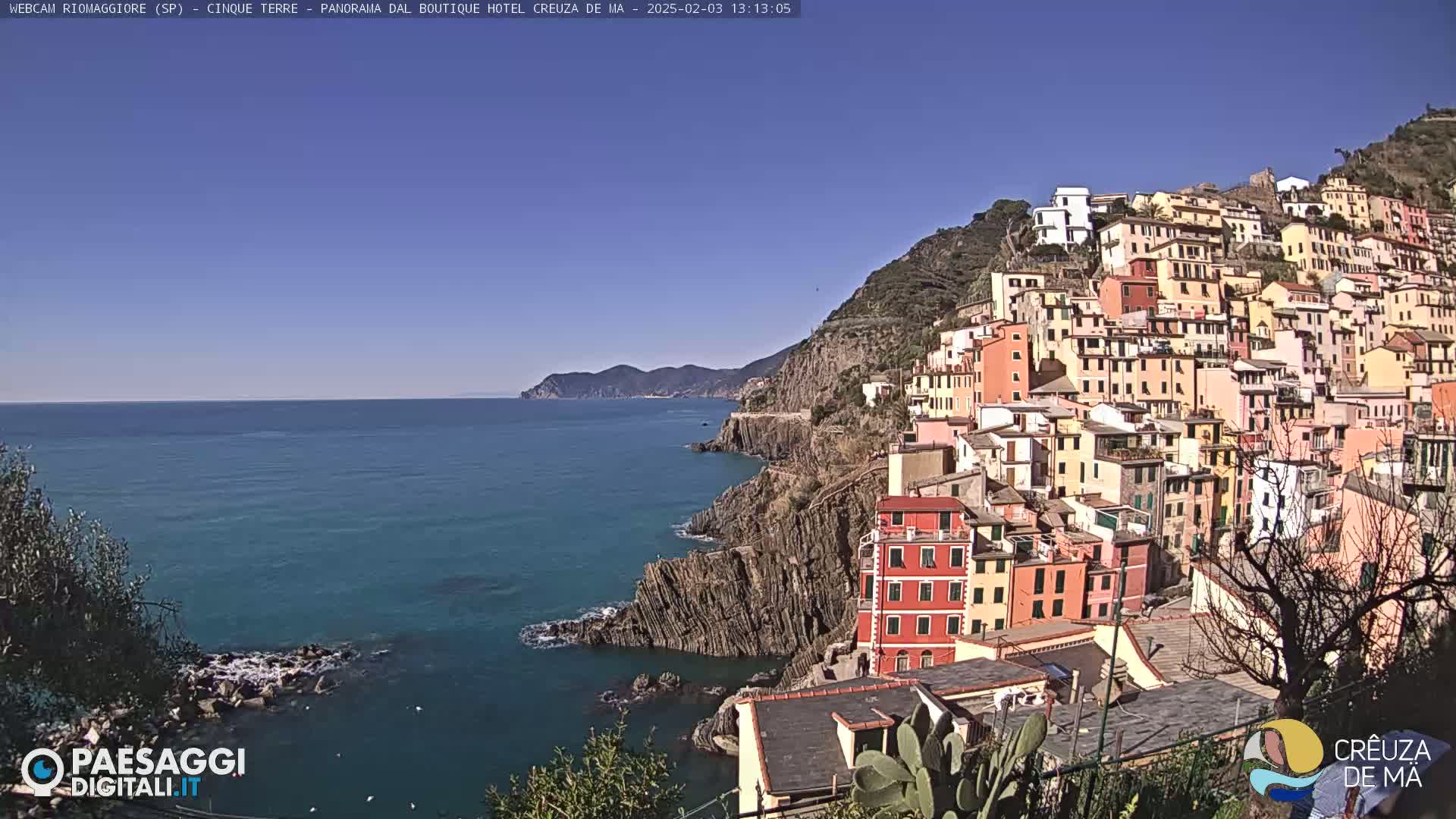 A colorful hillside village clings to a rocky coastline overlooking a calm blue sea under a clear blue sky.