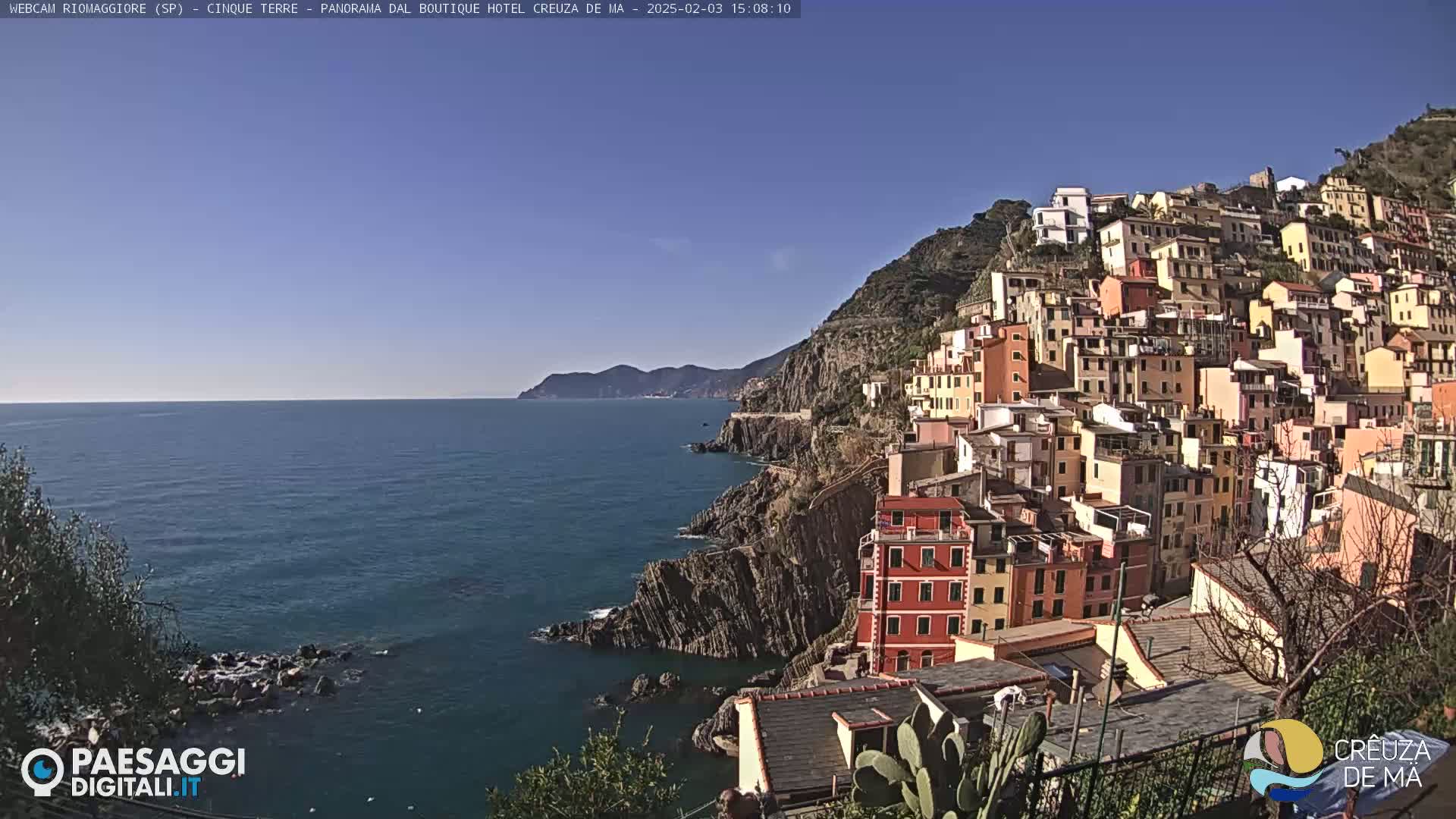 A colorful village clings to a steep hillside overlooking a calm, blue sea under a clear, sunny sky.