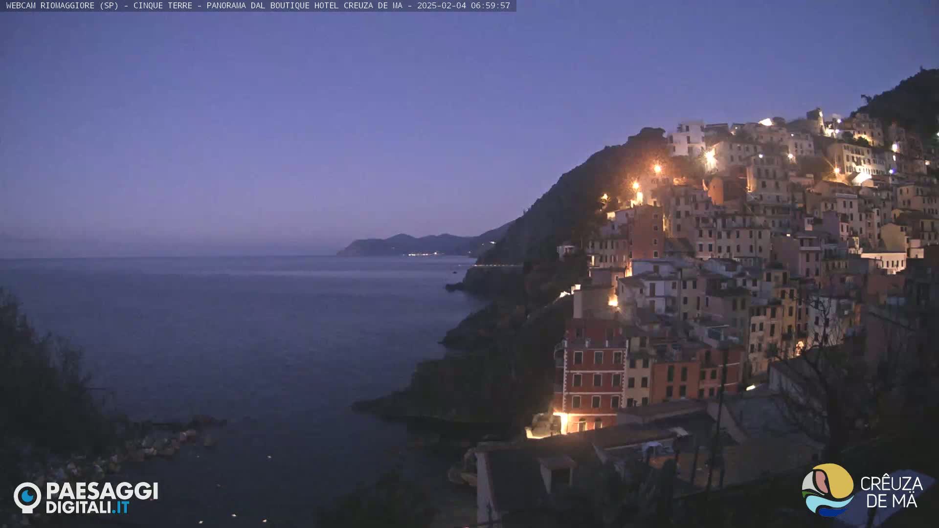 A coastal town illuminated at twilight, nestled on a hillside overlooking a dark, calm sea under a clear, dusky sky.