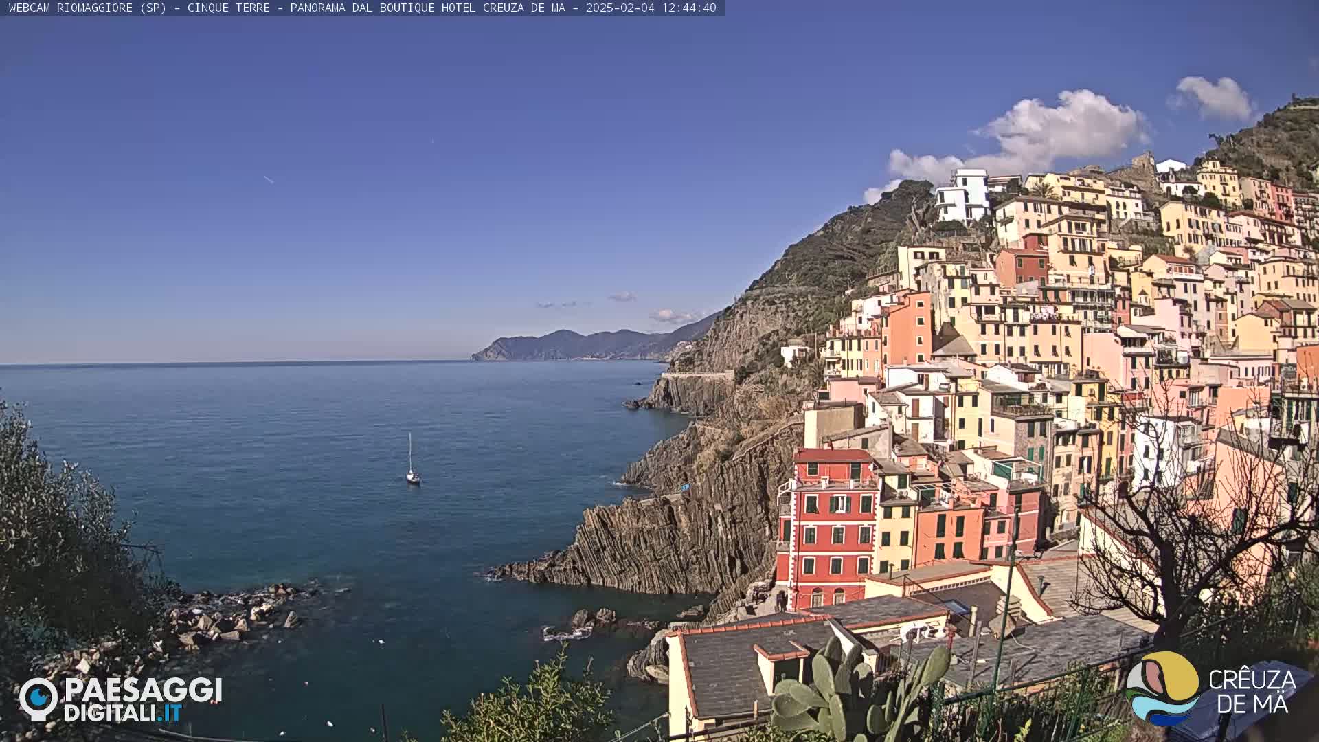 A coastal town of colorful buildings clings to a steep hillside overlooking a calm blue sea under a mostly sunny sky with a few scattered clouds.