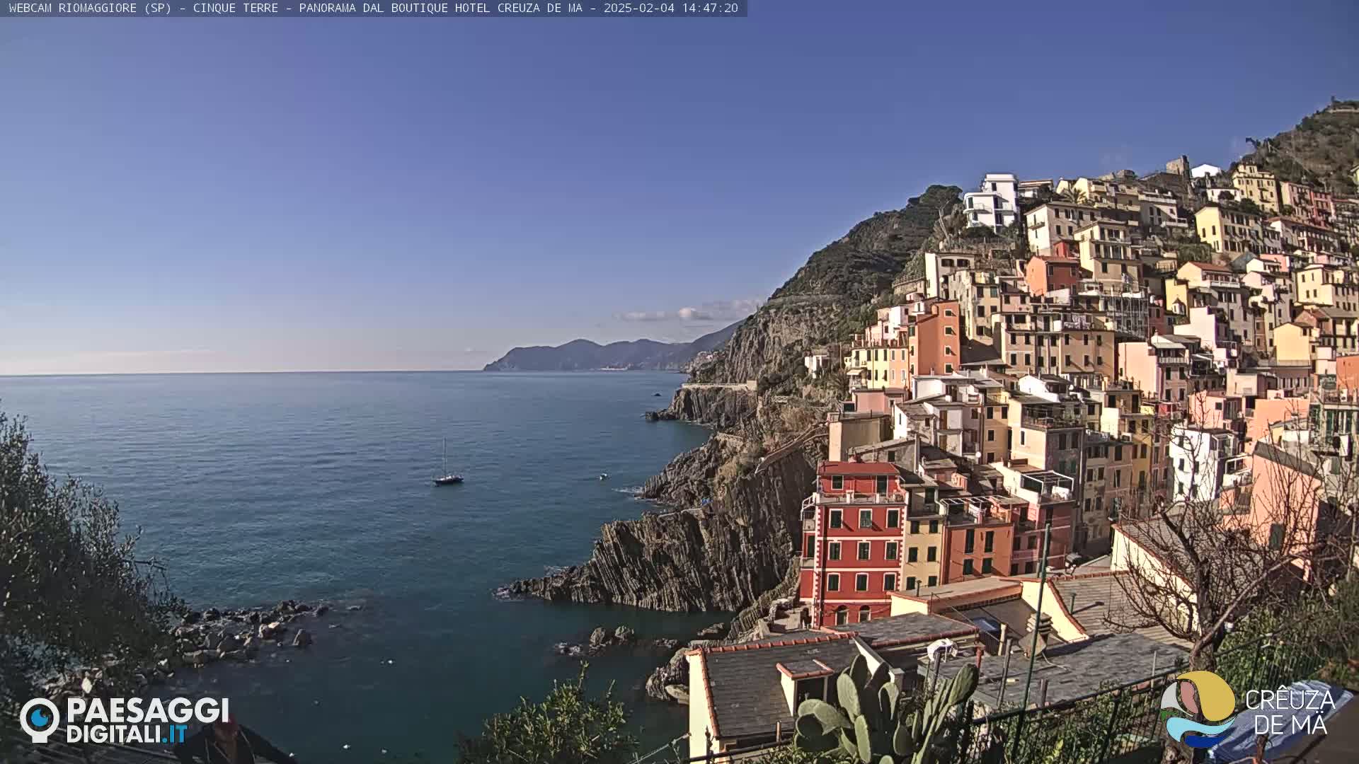 A colorful hillside village clings to a rocky coastline overlooking a calm, blue sea under a clear, sunny sky.