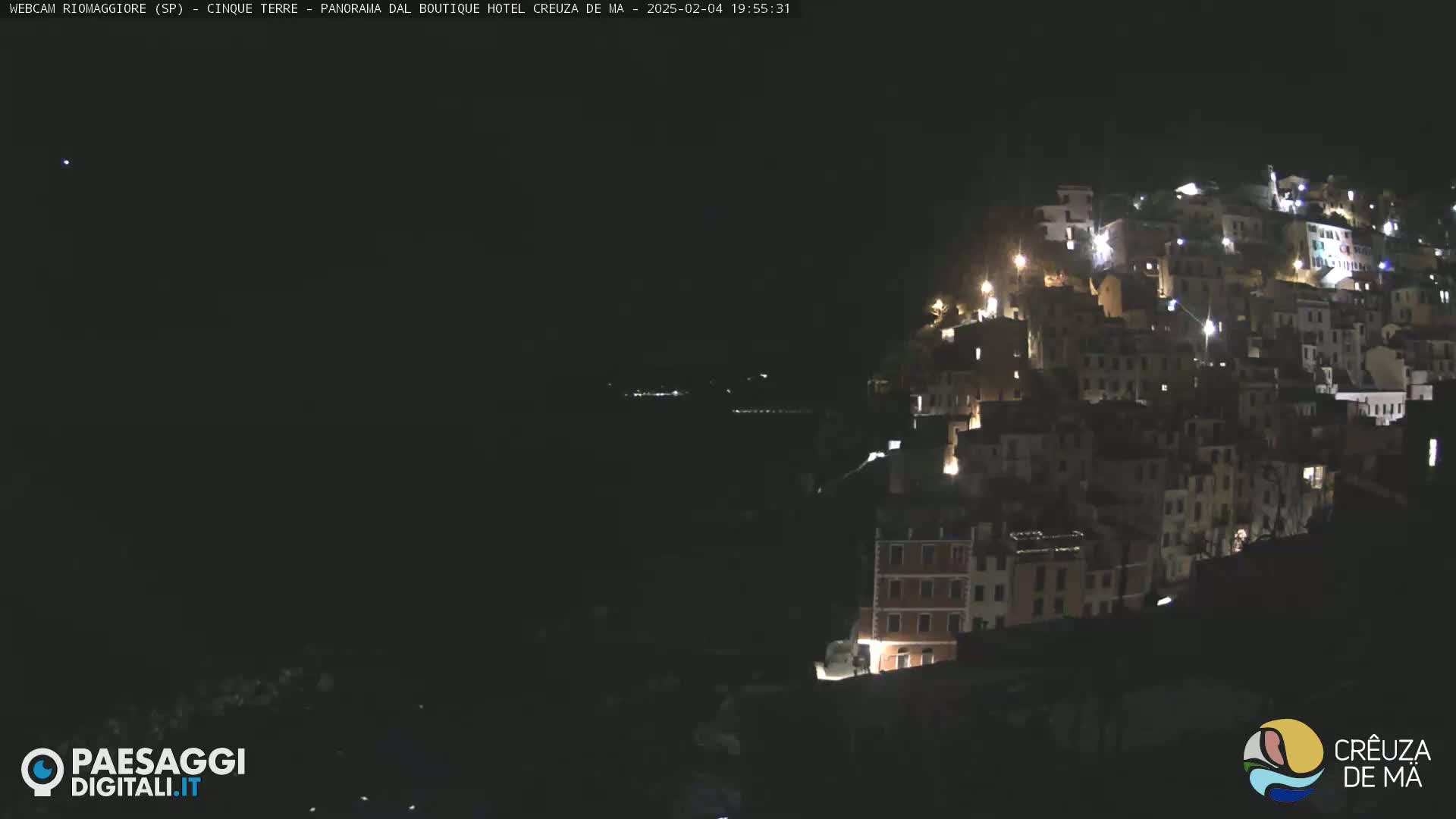 A nighttime view of a hillside town illuminated by lights, overlooking a dark body of water under a clear, dark sky.