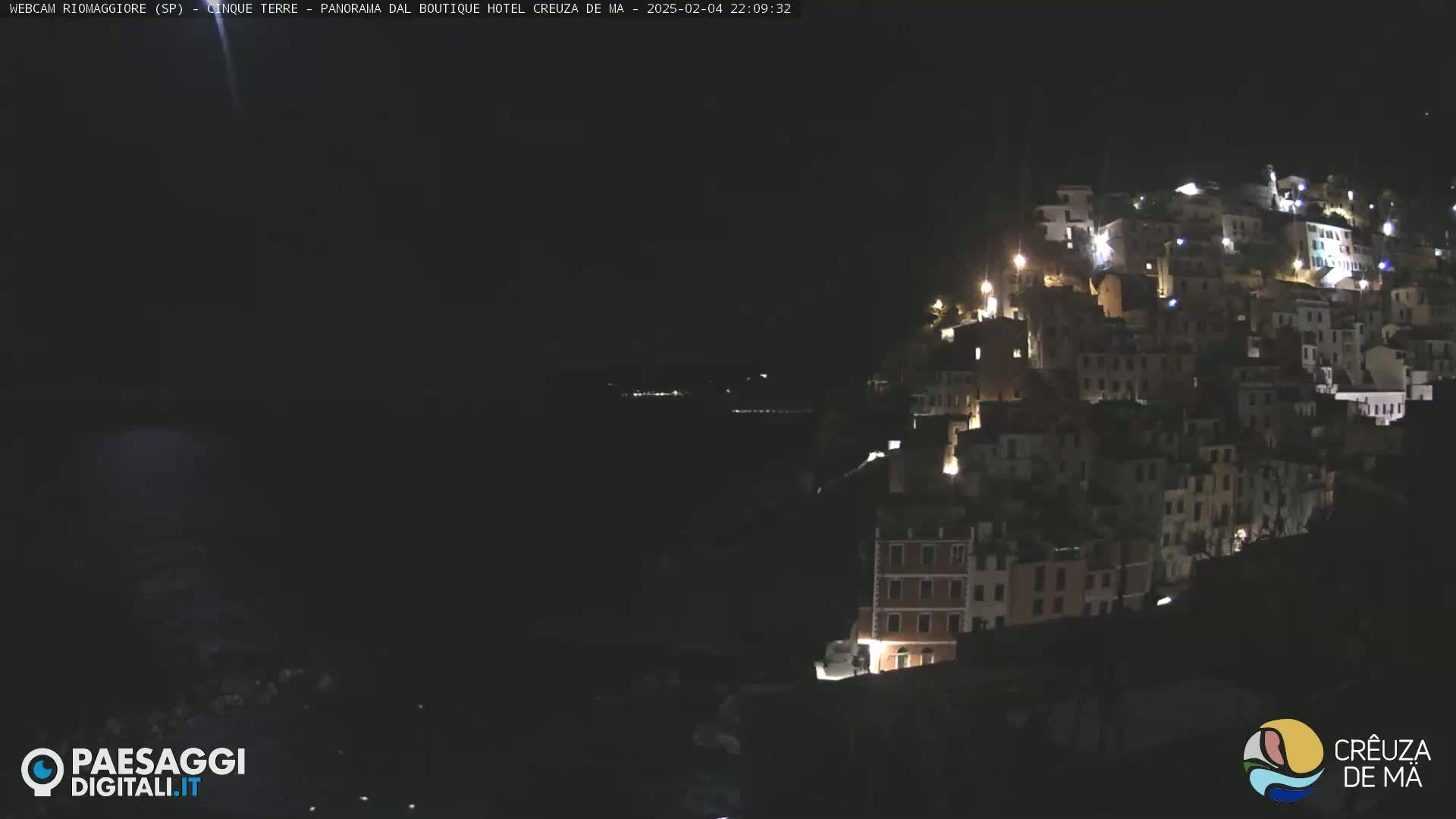 A nighttime view of a hillside town with many lit windows, overlooking a dark body of water.