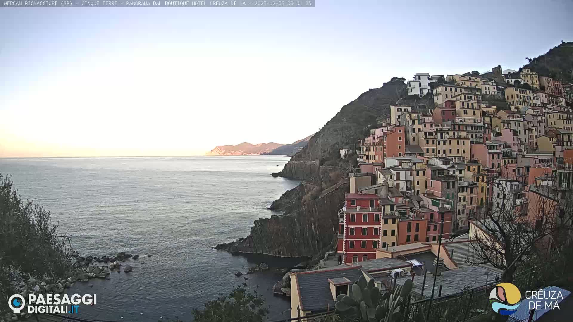 A colorful hillside village clings to a rocky coastline overlooking a calm sea under a clear, light sky.