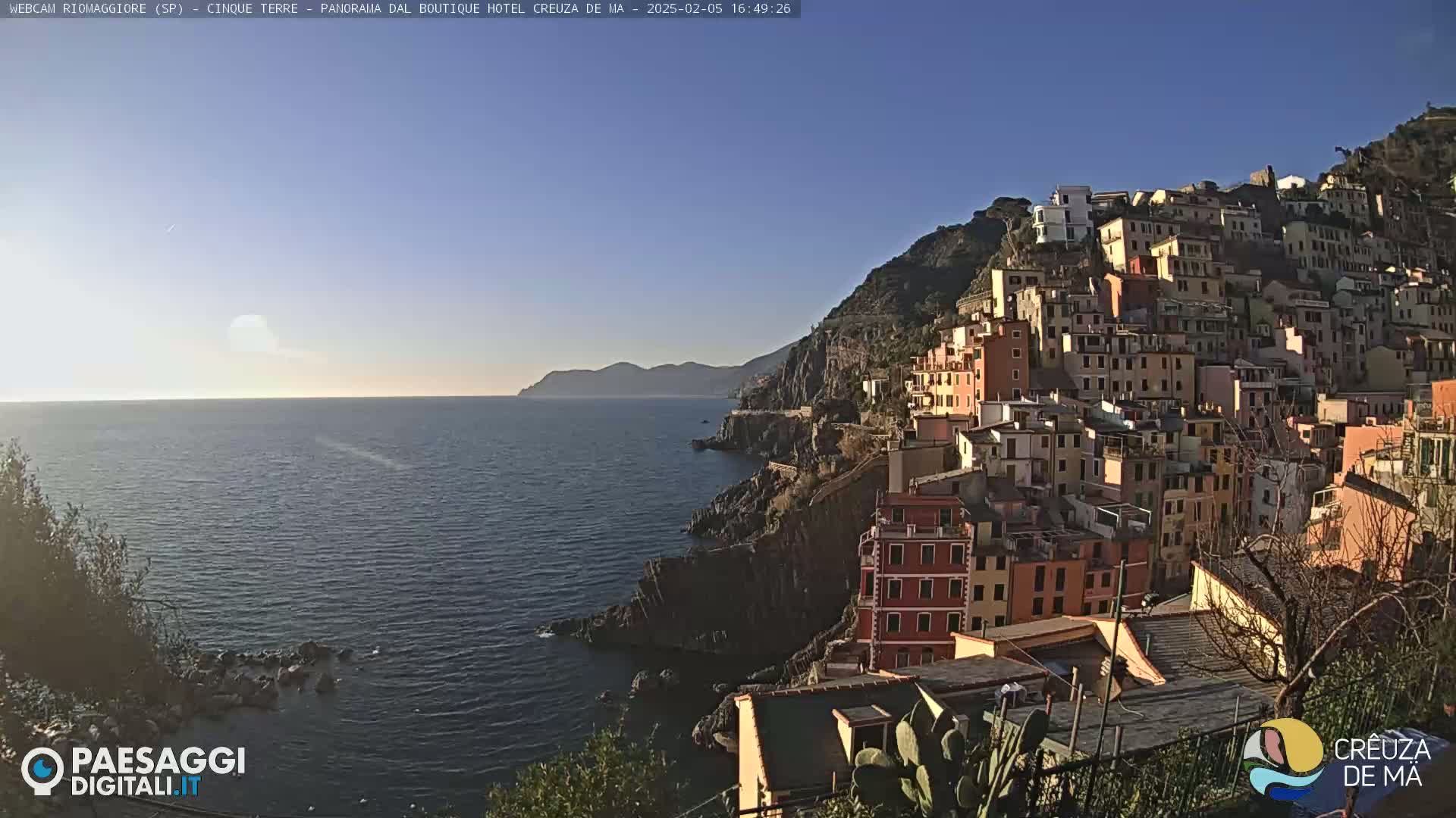 A coastal town built on a steep hillside overlooks a calm, blue sea under a clear, bright sky.
