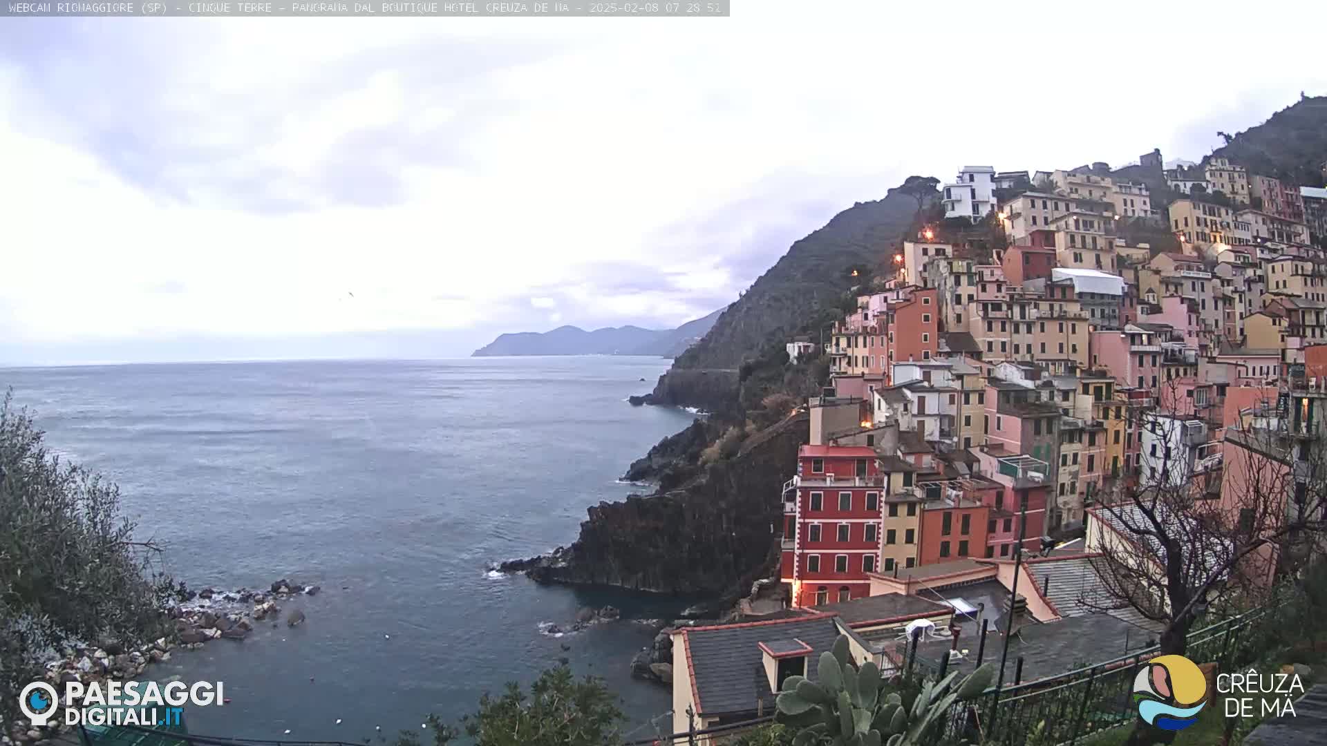 A coastal town of colorful buildings climbs a steep hillside overlooking a calm sea under a cloudy sky.