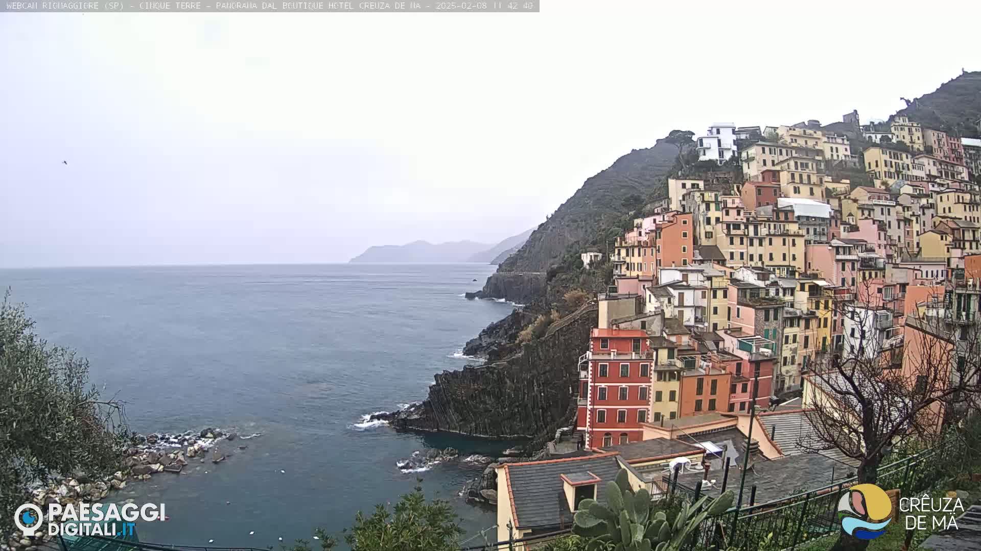 A coastal town with colorful buildings clinging to a steep cliff overlooking a calm, gray sea under an overcast sky.
