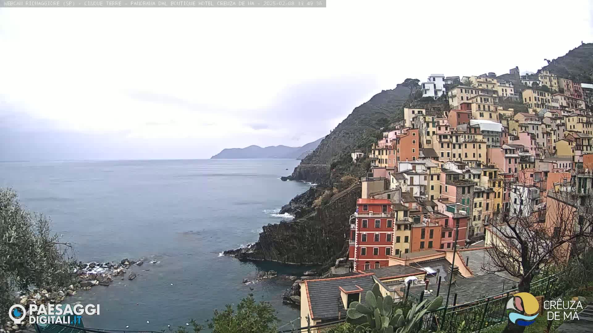 A coastal town of colorful buildings clings to a steep hillside overlooking a calm sea under an overcast sky.