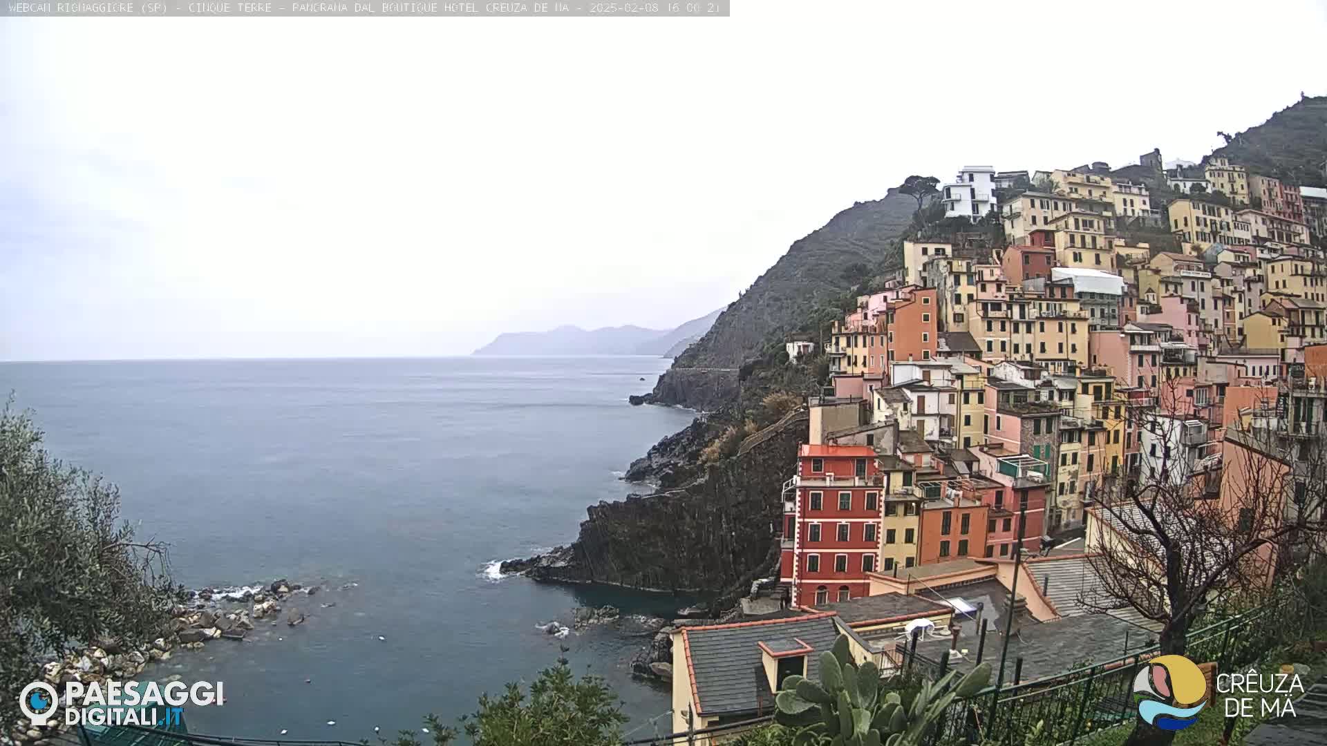 A colorful hillside village clings to a rocky coast overlooking a calm, gray sea under an overcast sky.