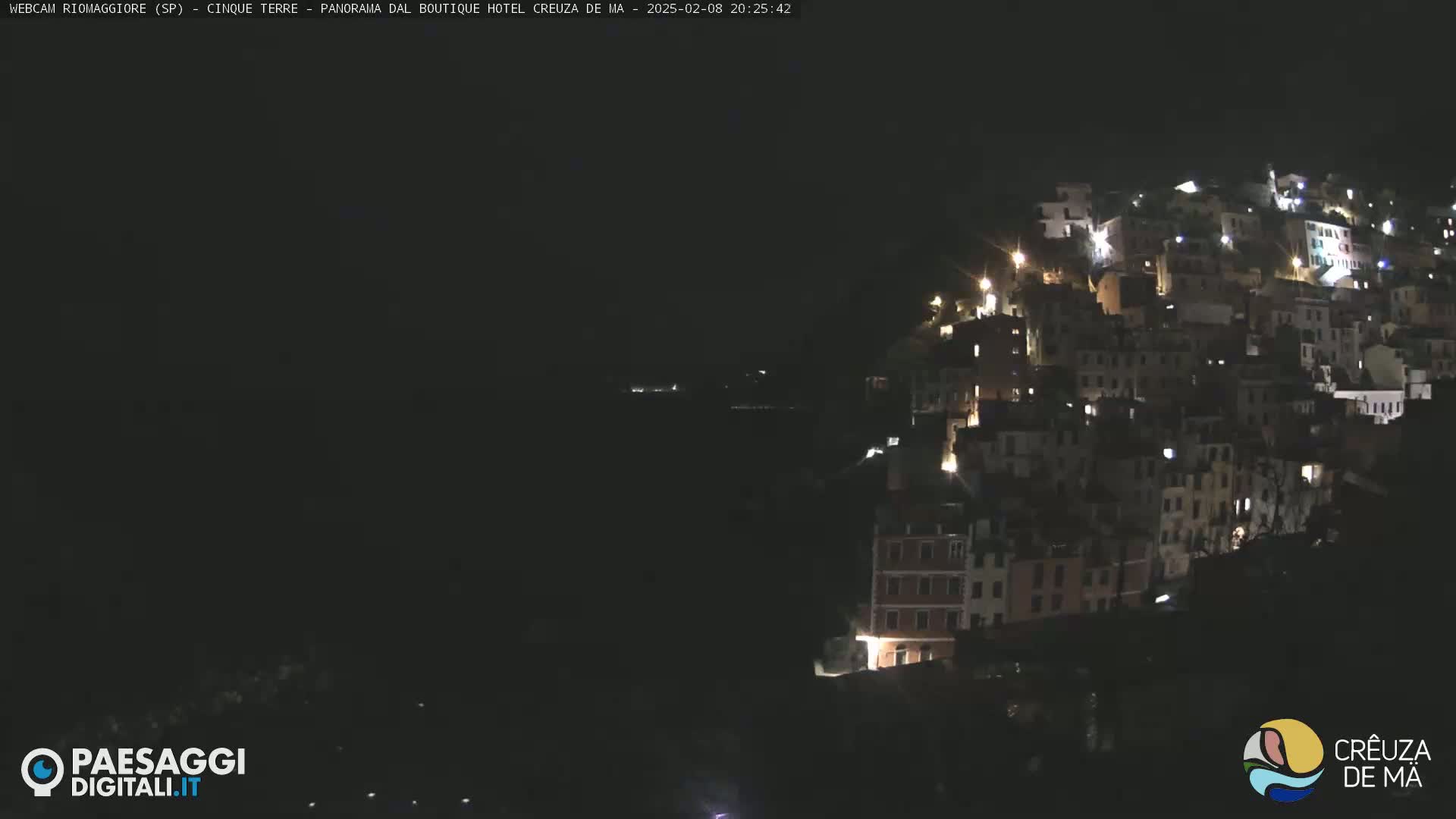 A nighttime view of a hillside town with many illuminated buildings, overlooking a dark body of water.