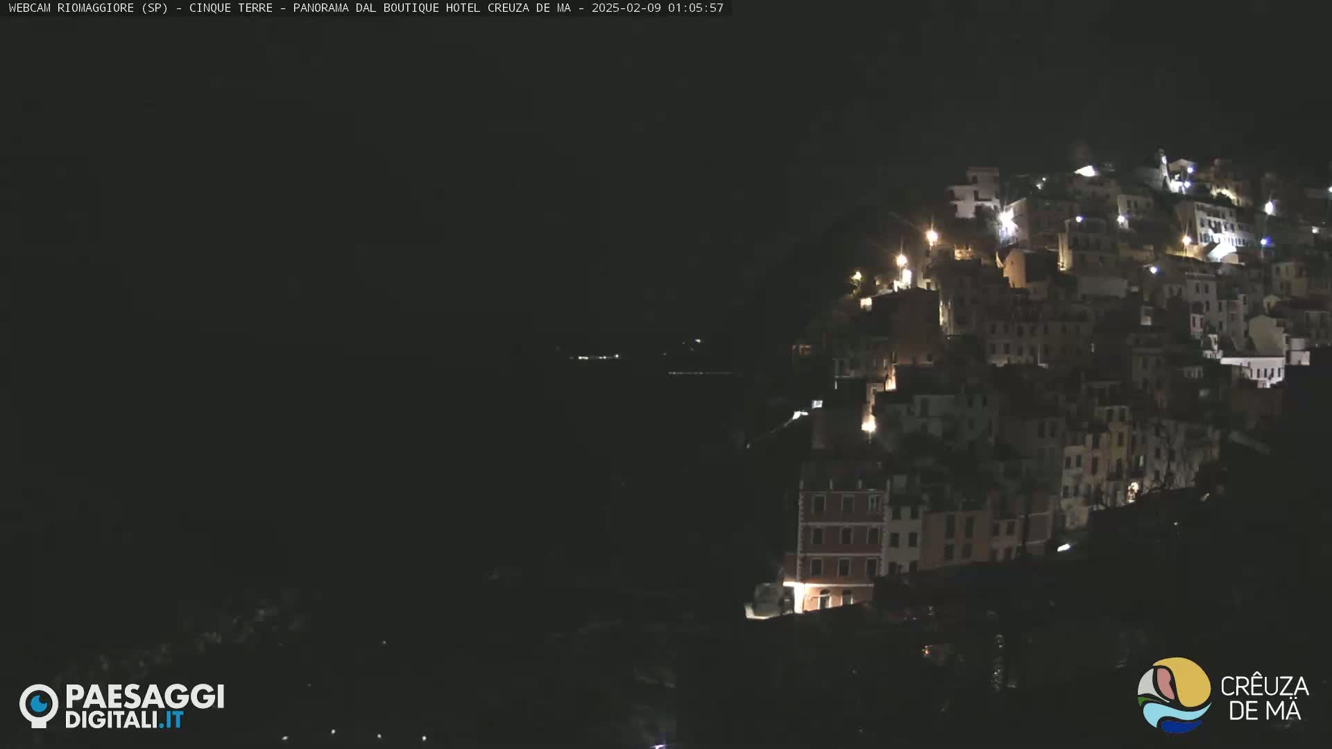 A nighttime view of a hillside town illuminated by scattered lights, overlooking a dark body of water under a clear, dark sky.