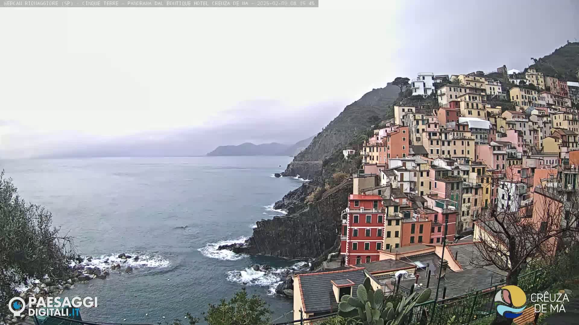 A coastal village of colorful buildings clings to a steep hillside overlooking a choppy sea under an overcast sky.