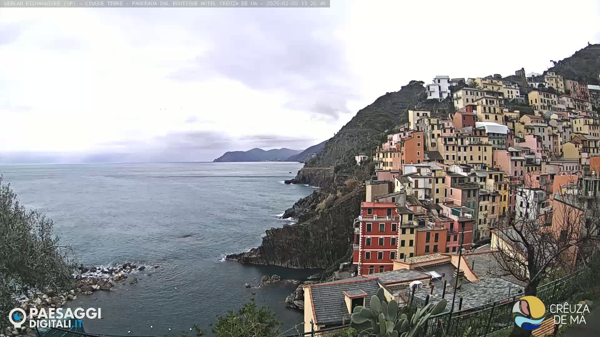 A coastal village of colorful buildings clings to a steep hillside overlooking a calm sea under a cloudy sky.