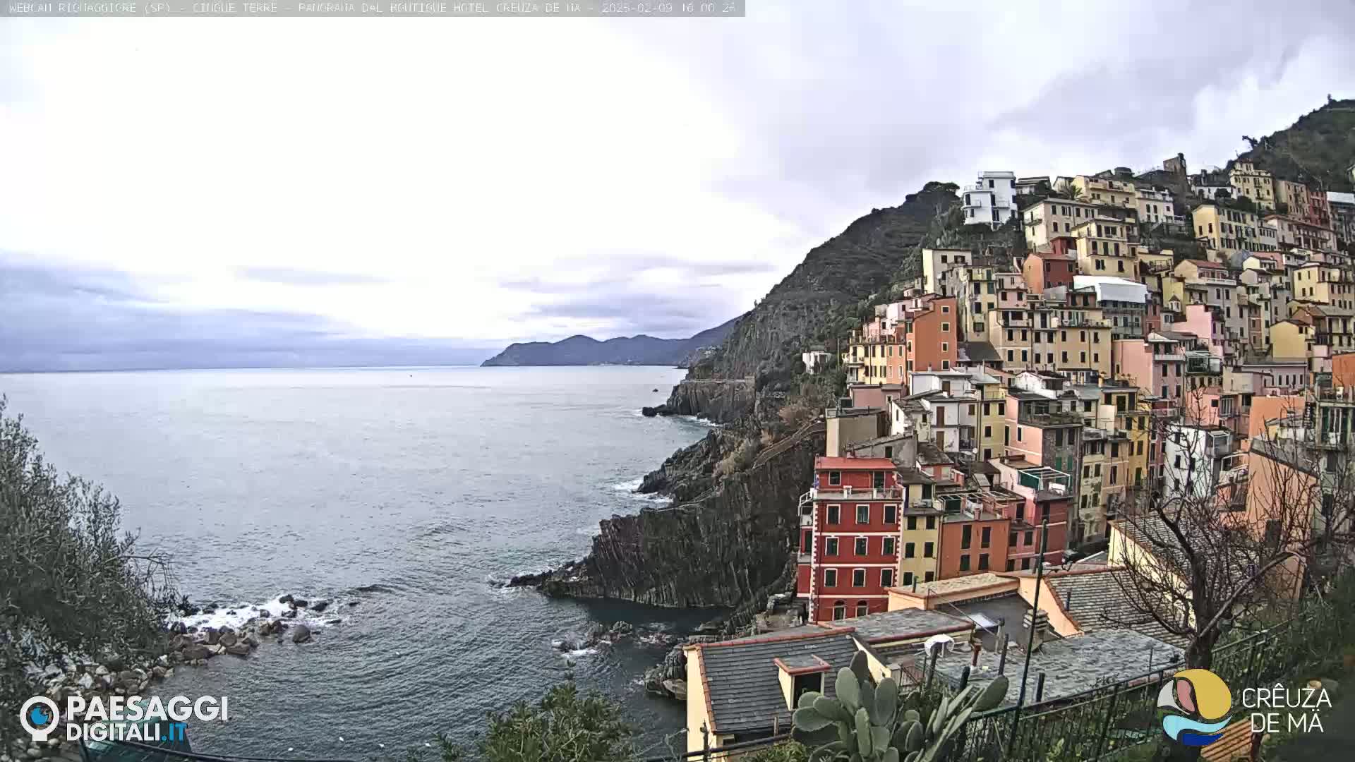 A coastal town with colorful buildings clinging to a steep cliffside overlooks a calm sea under a cloudy sky.
