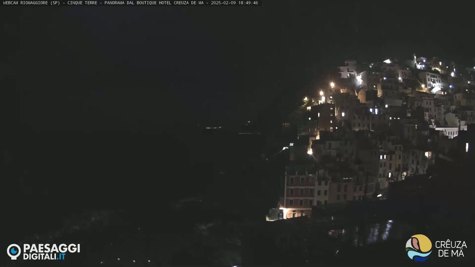 A nighttime view of a hillside town illuminated by scattered lights, overlooking a dark body of water.