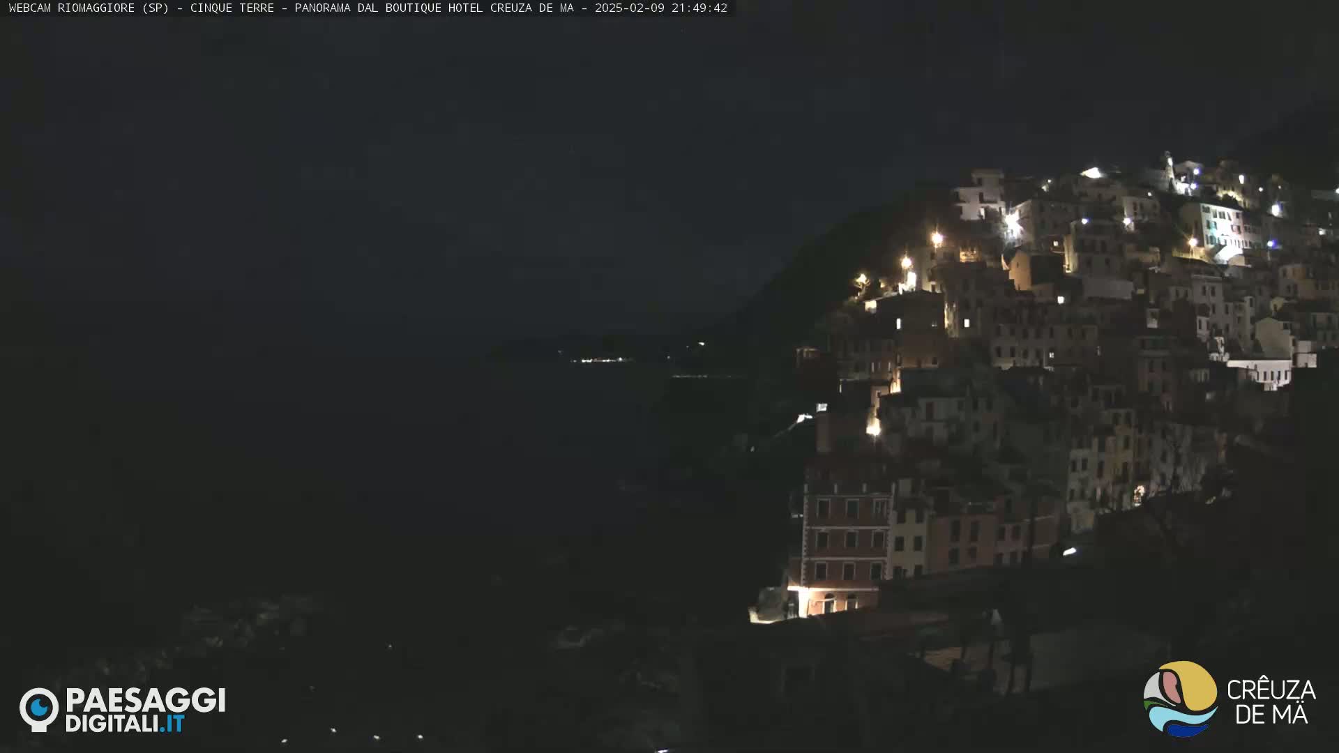 A nighttime view of a hillside village with many lit windows overlooking a dark body of water.