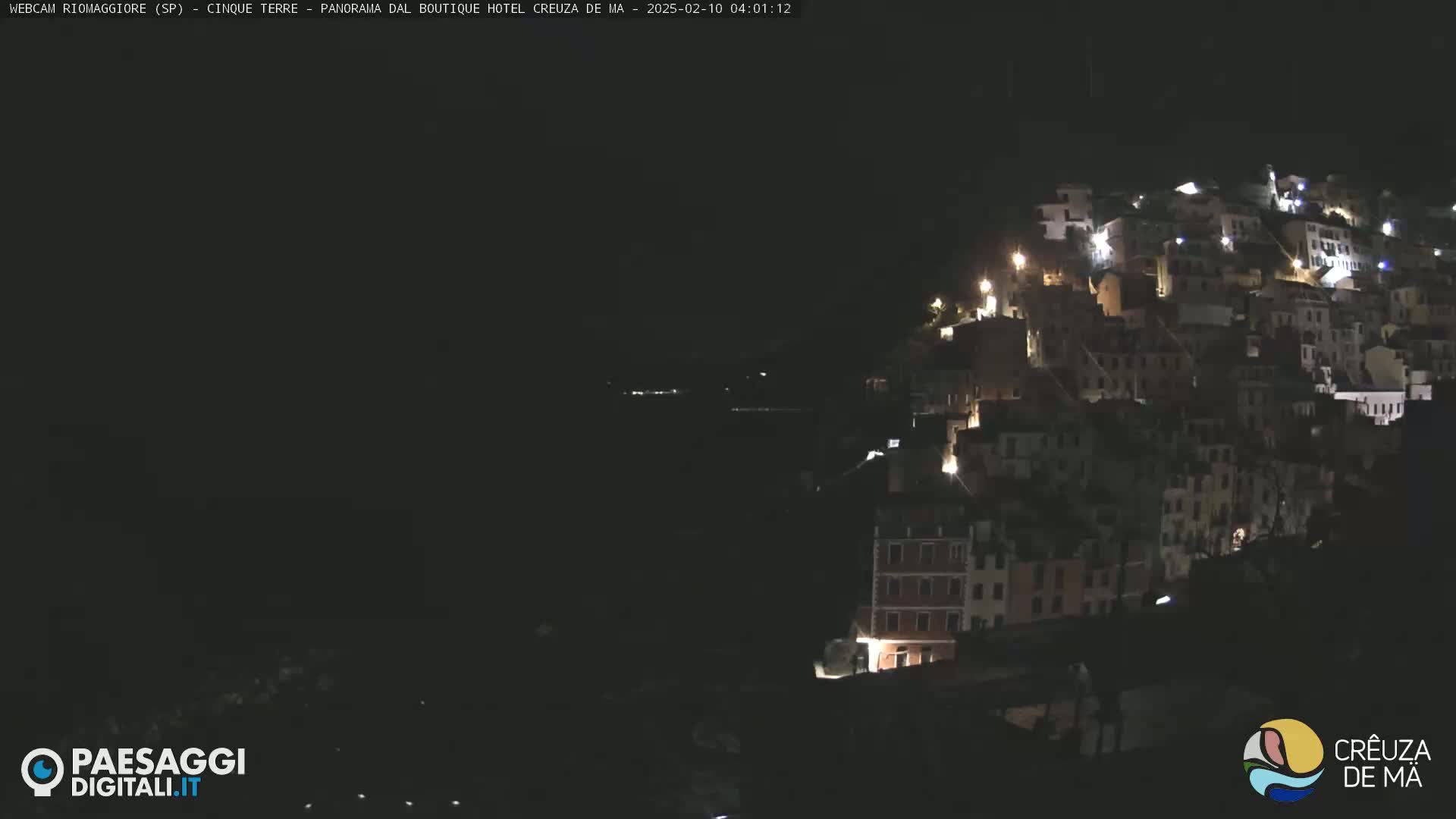 A nighttime view of a hillside town illuminated by lights, overlooking a dark body of water.
