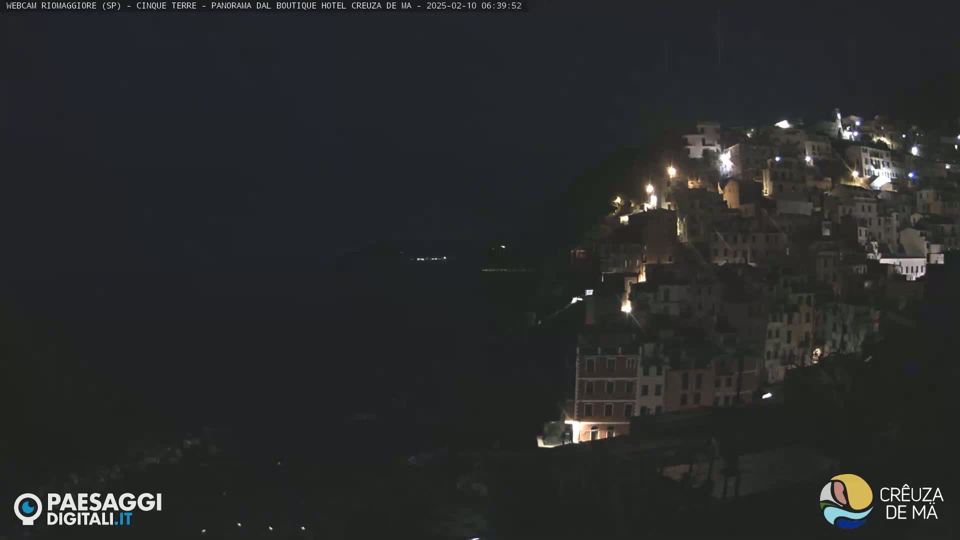 A nighttime view of a hillside town illuminated by lights, overlooking a dark body of water under a clear, dark sky.