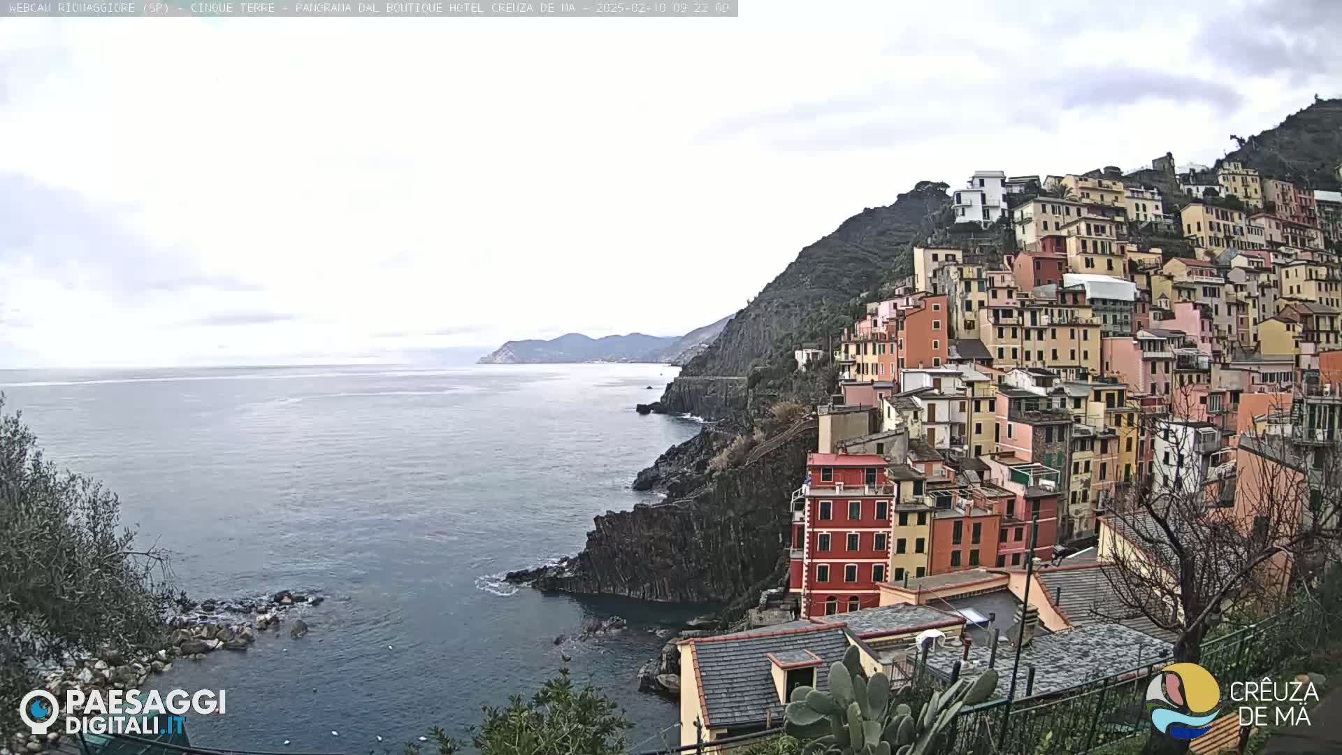 A coastal village of colorful buildings clings to a steep hillside overlooking a calm sea under a cloudy sky.