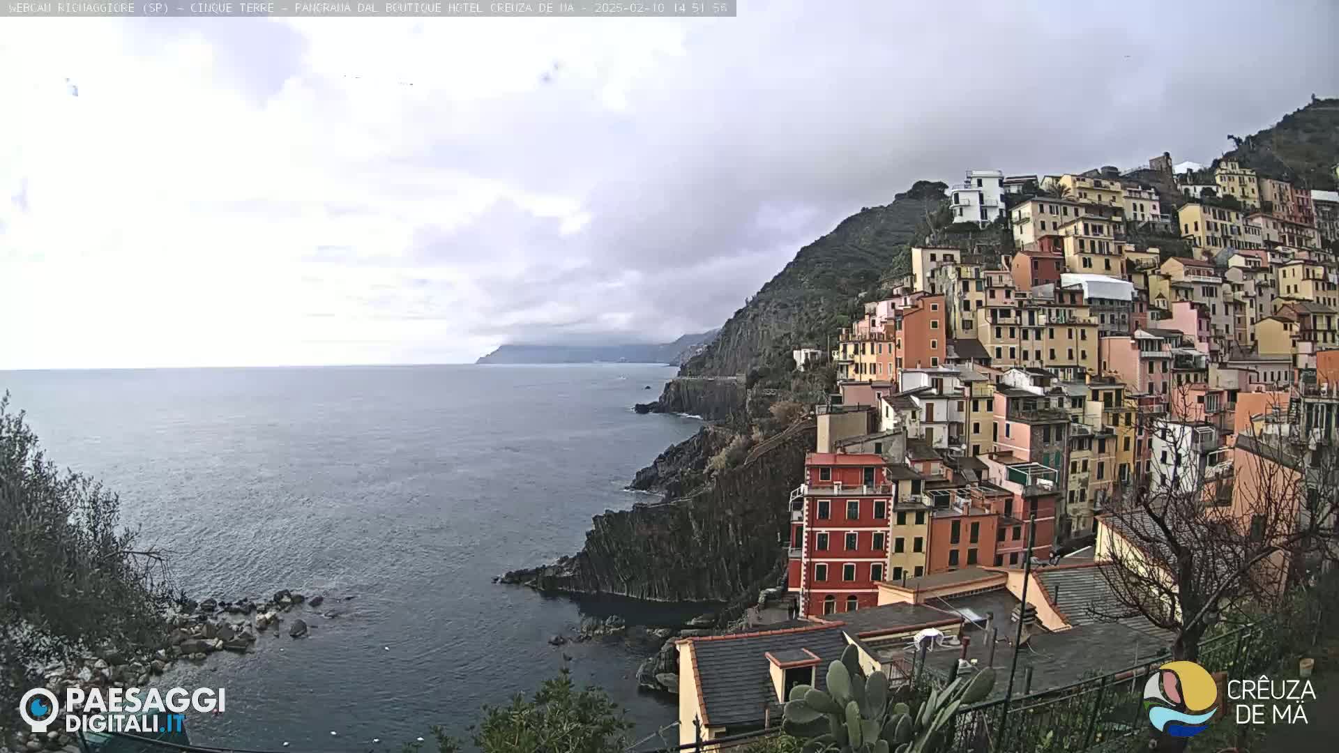 A colorful village clings to a steep, rocky hillside overlooking a calm, overcast ocean.