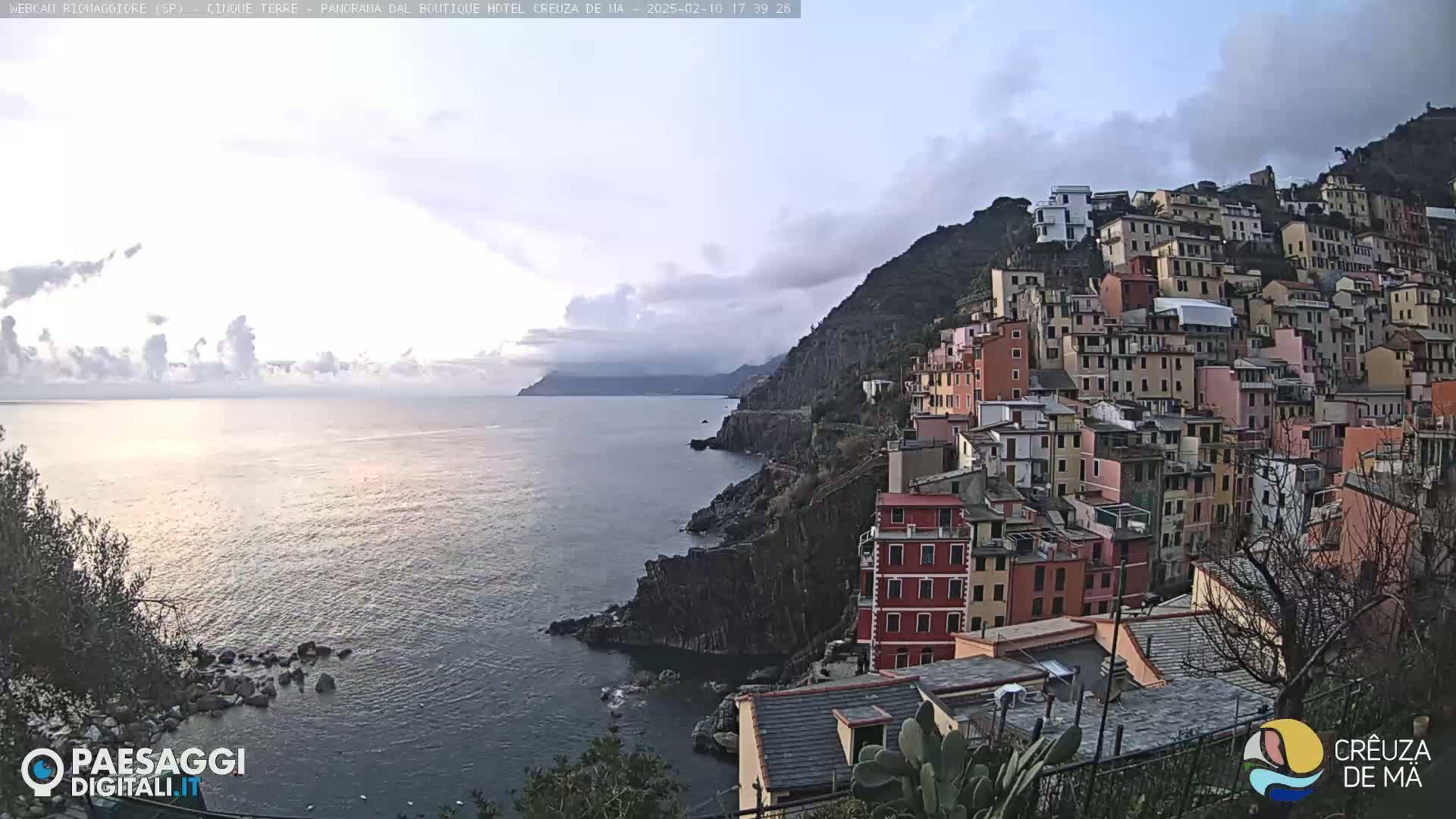 A coastal town with colorful buildings clinging to a steep hillside overlooks a calm sea under a partly cloudy sky.