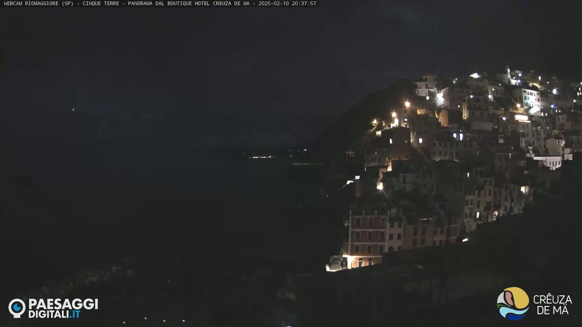 A nighttime view of a hillside town illuminated by lights, overlooking a dark body of water under a clear sky.