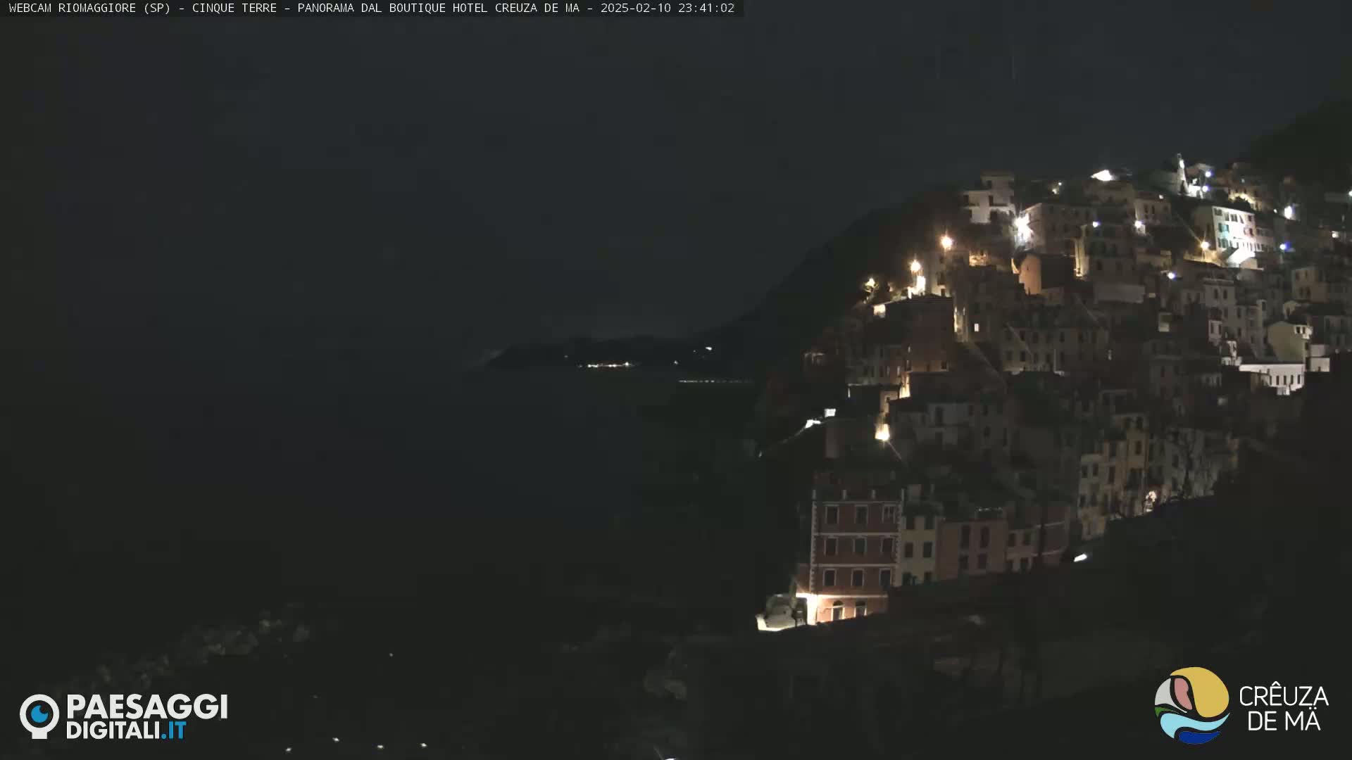 A nighttime coastal village with illuminated buildings clings to a dark hillside overlooking a black sea under a dark sky.