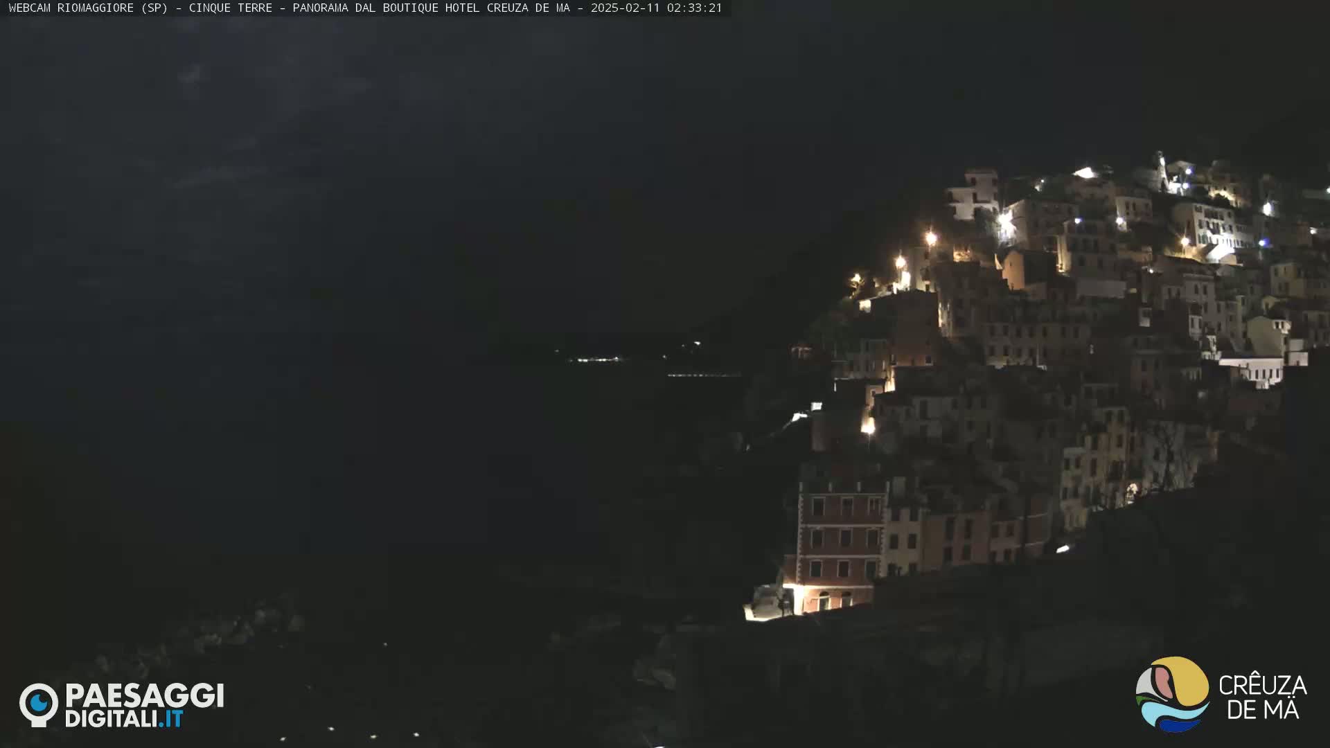 A nighttime view of a hillside town illuminated by lights, overlooking a dark body of water under a mostly clear sky.