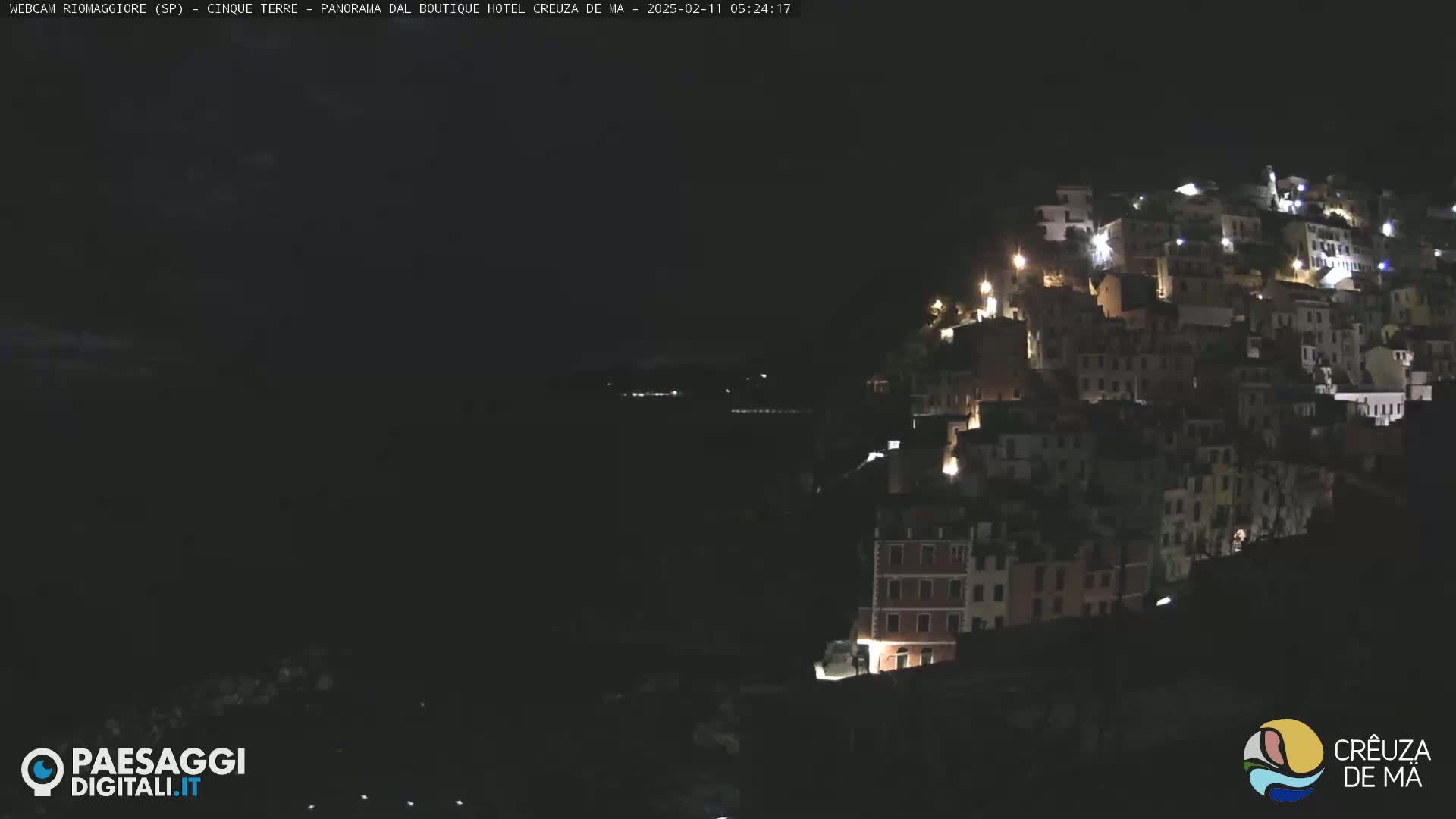 A nighttime view of a hillside town illuminated by scattered lights, overlooking a dark body of water under a clear, dark sky.