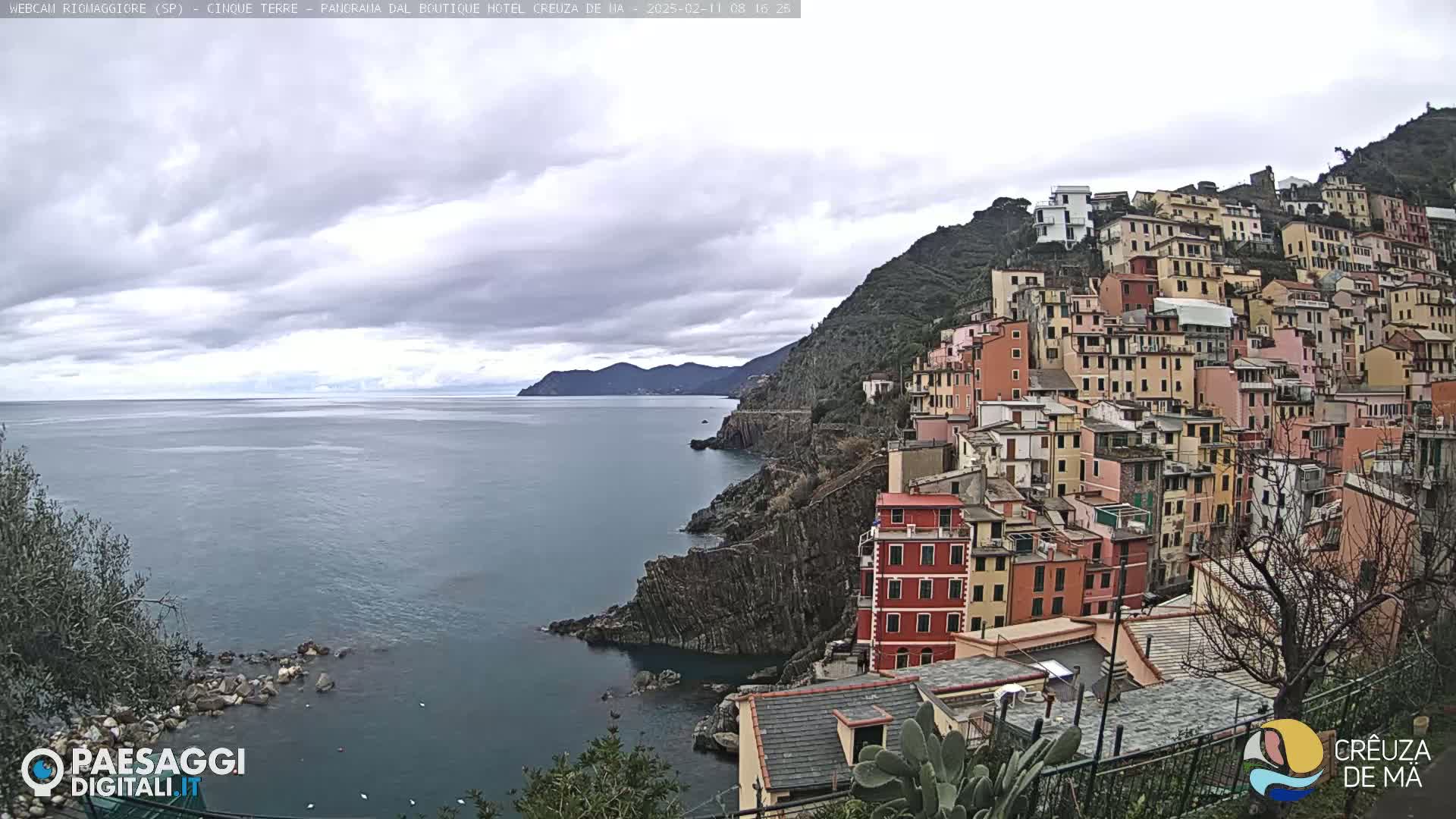 A coastal village of colorful buildings climbs a steep hillside overlooking a calm sea under a cloudy sky.