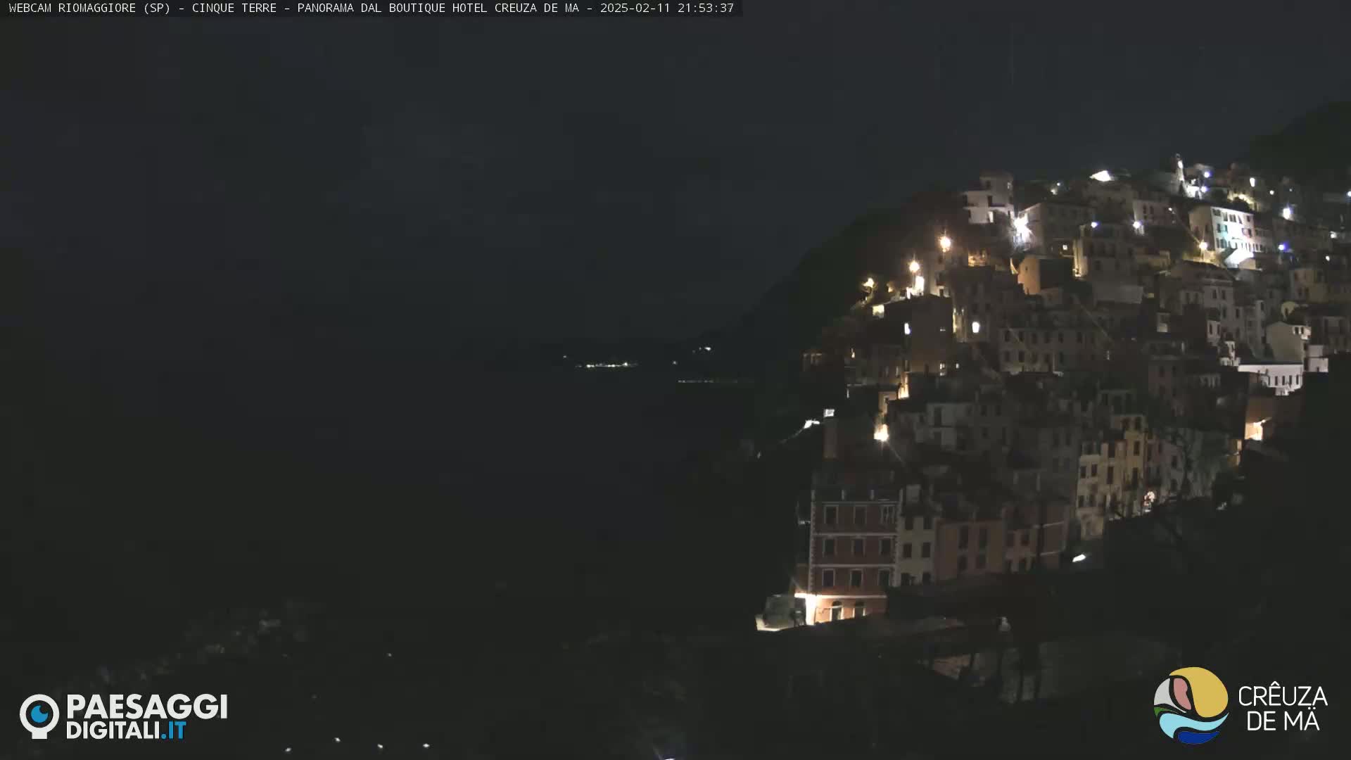 A nighttime view of a hillside town illuminated by scattered lights, overlooking a dark body of water under a clear, dark sky.