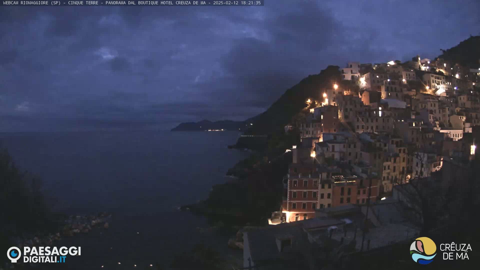 A coastal town at night with its buildings illuminated, sits on a hillside overlooking a dark sea under a cloudy sky.